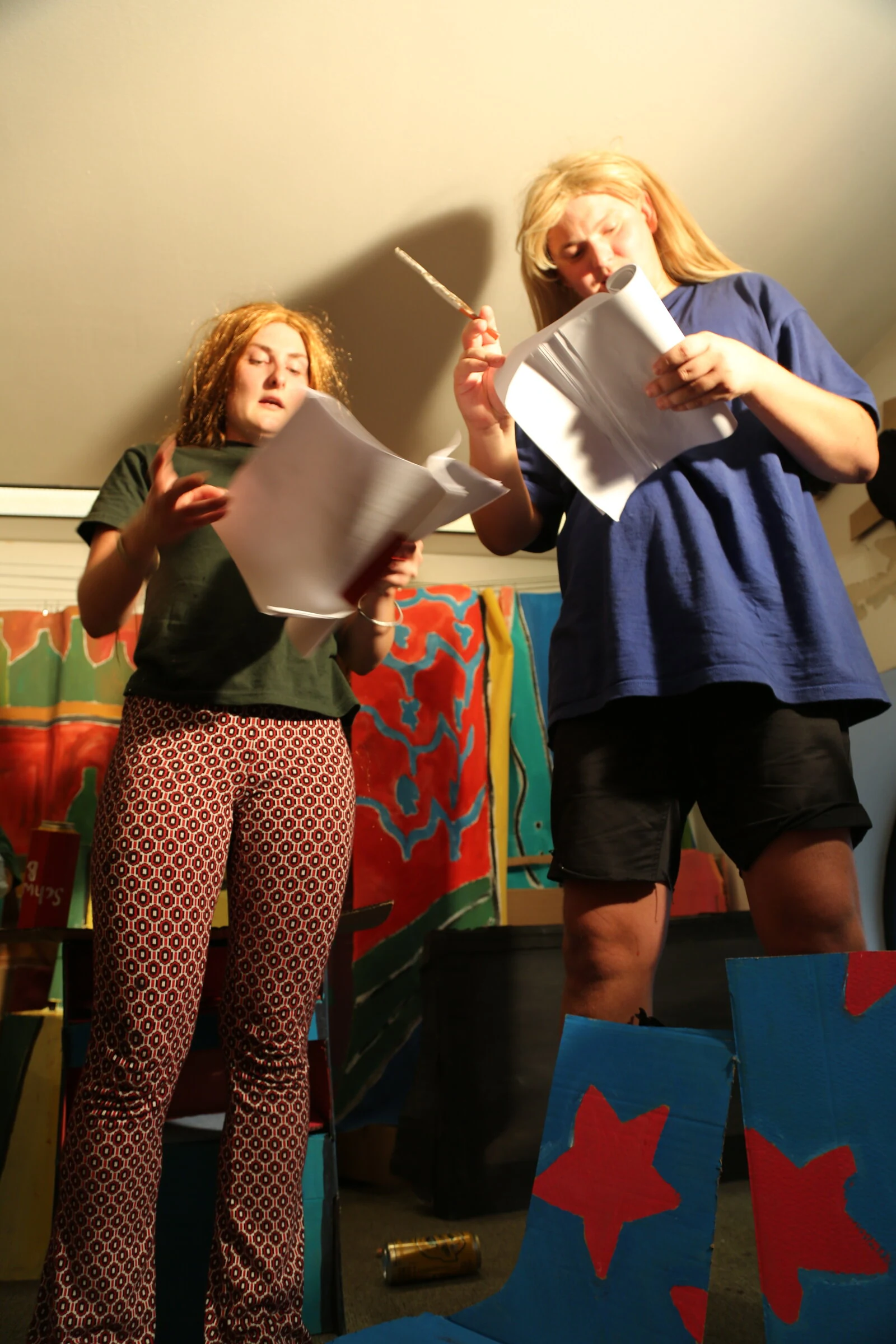 Two performers in green and blue shirts reading scripts beside the painted backdrop.