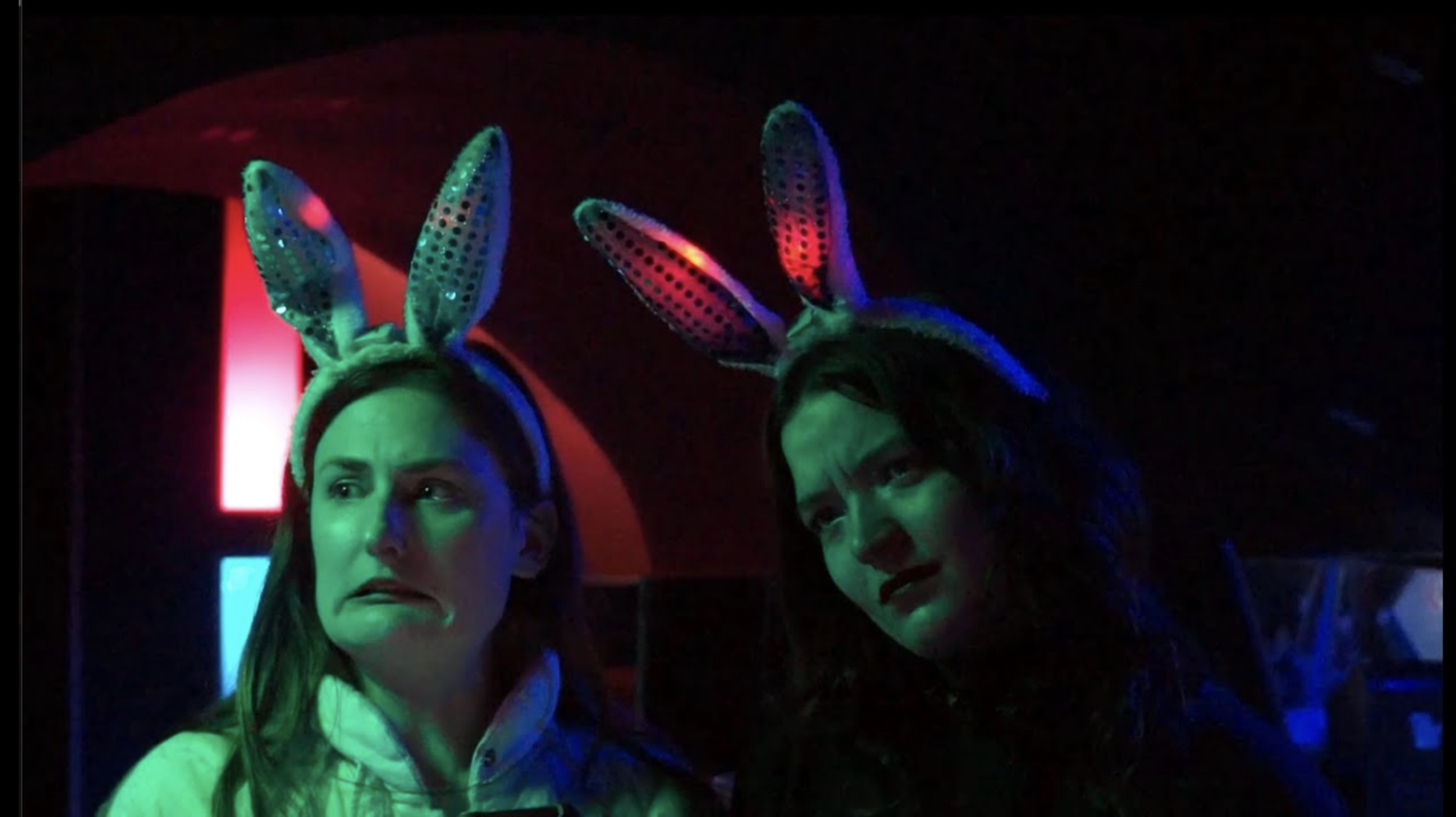 Two women wearing bunny-ear headbands under green, blue and pink party lighting.