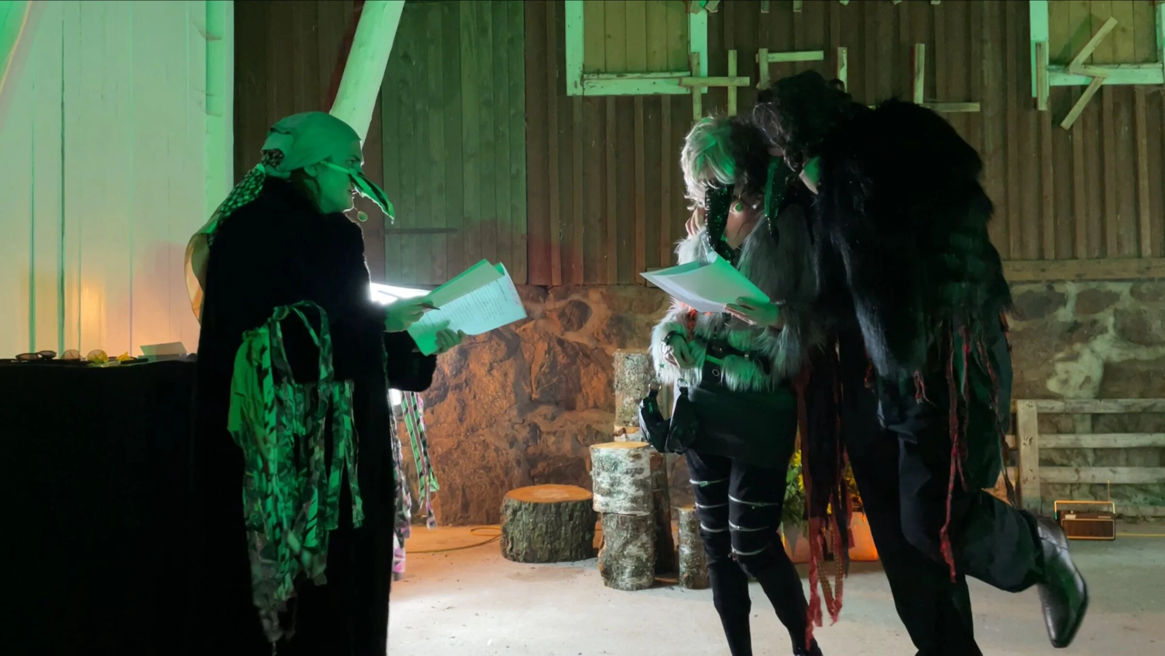 Three masked performers reading scripts together under green lighting.