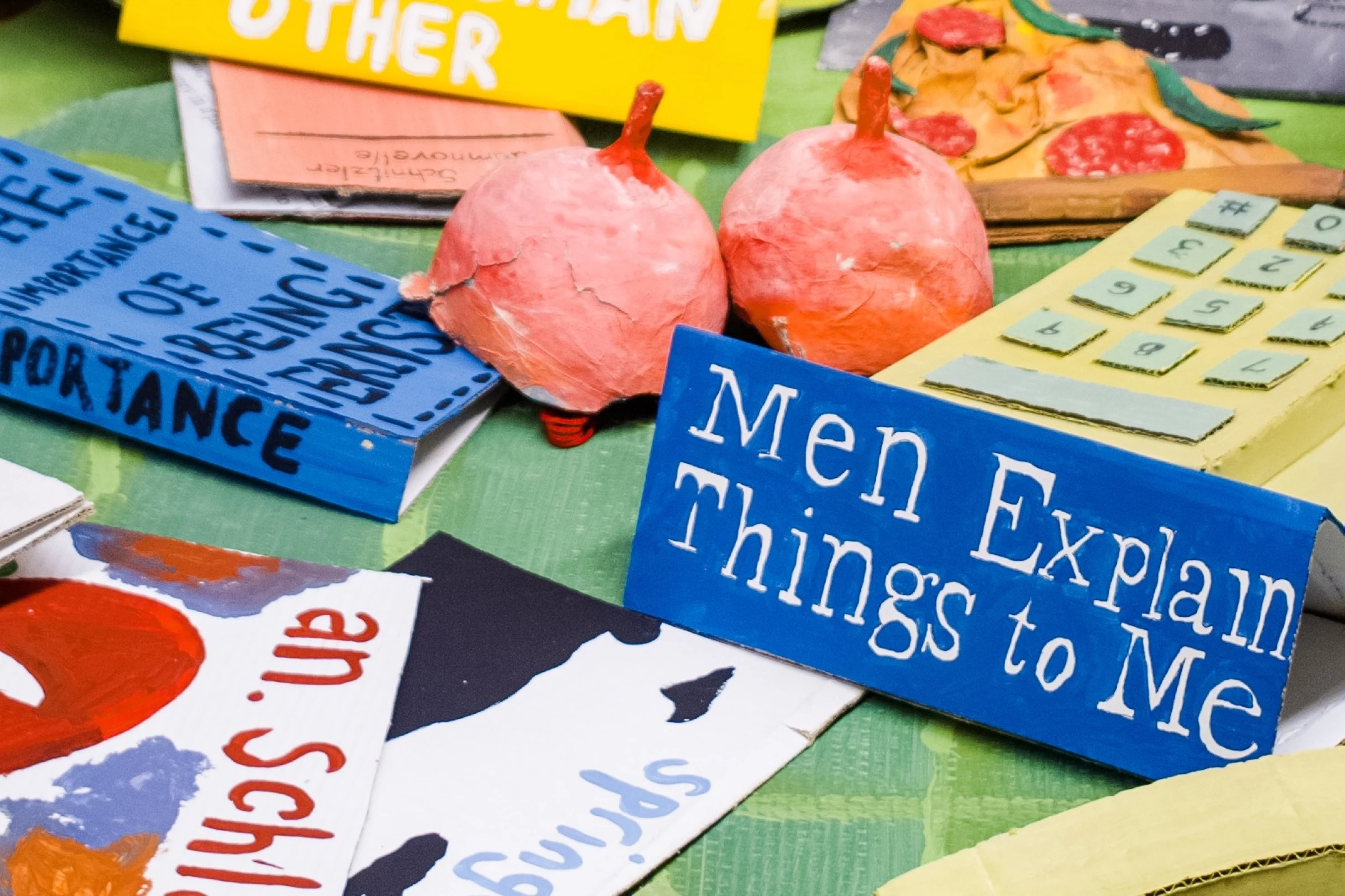A detail of the cardboard Men Explain Things to Me book beside pink sculptures and a yellow telephone.