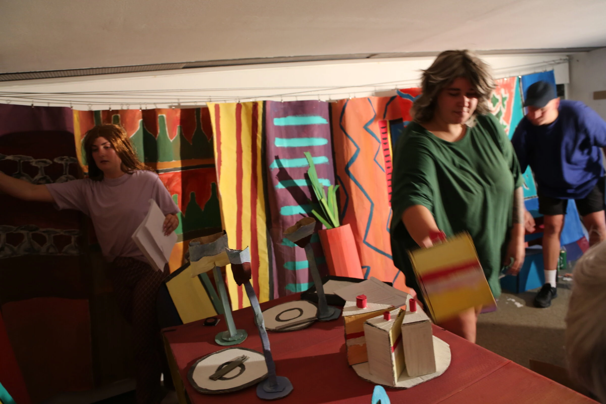 A performer in green moving a cardboard cake slice while two other performers stand nearby.