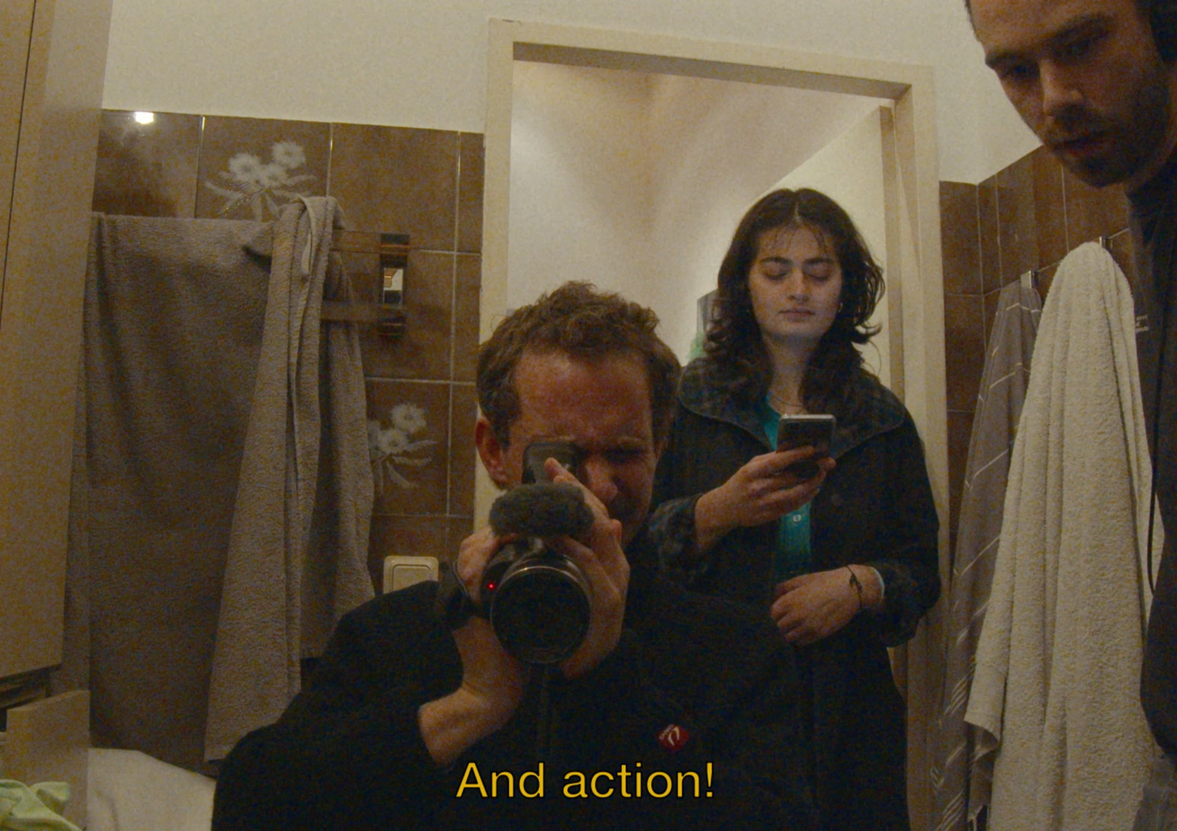 A camera operator and two people reflected in a bathroom mirror.