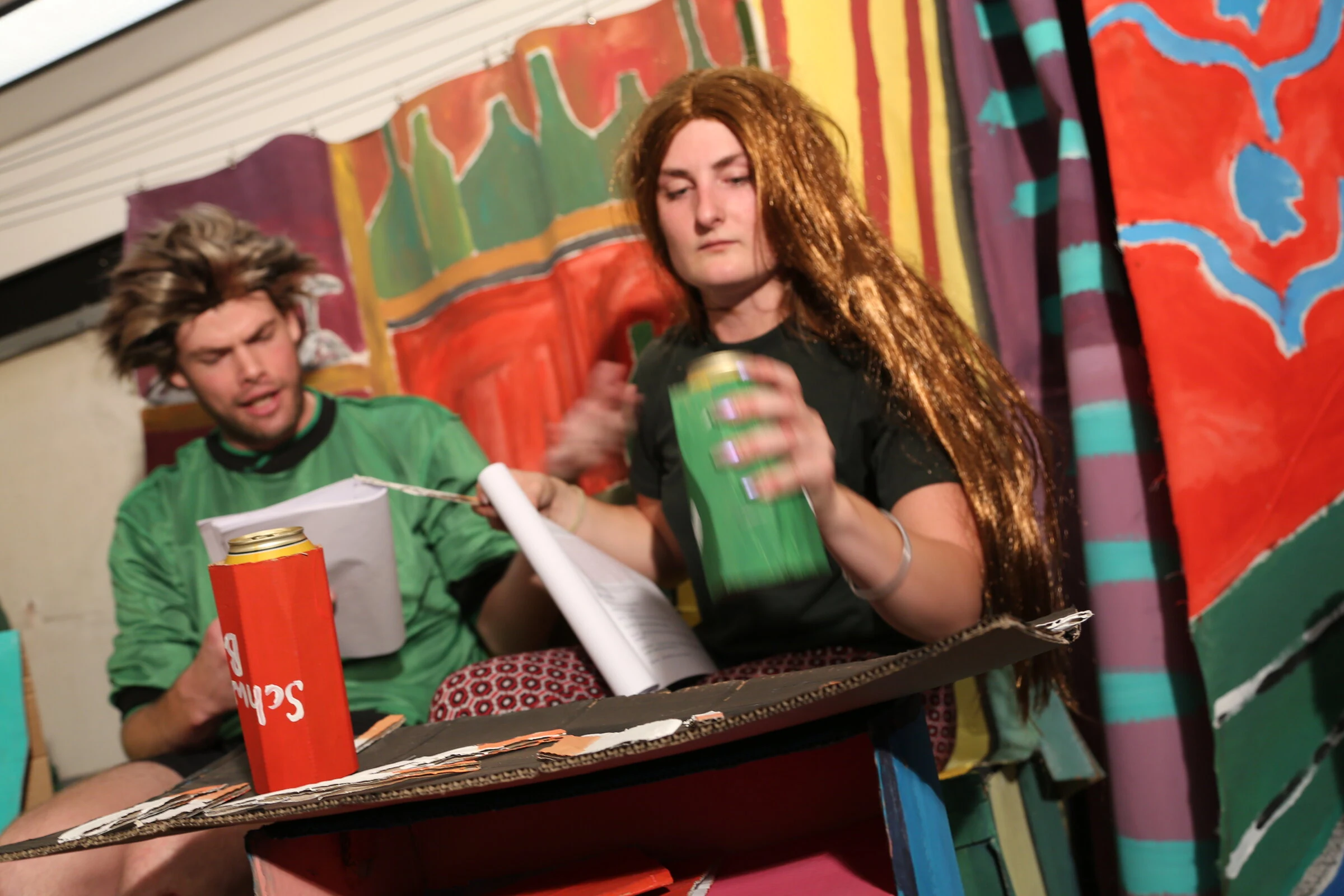 Two performers reading scripts at a cardboard table, one holding a green drink holder.