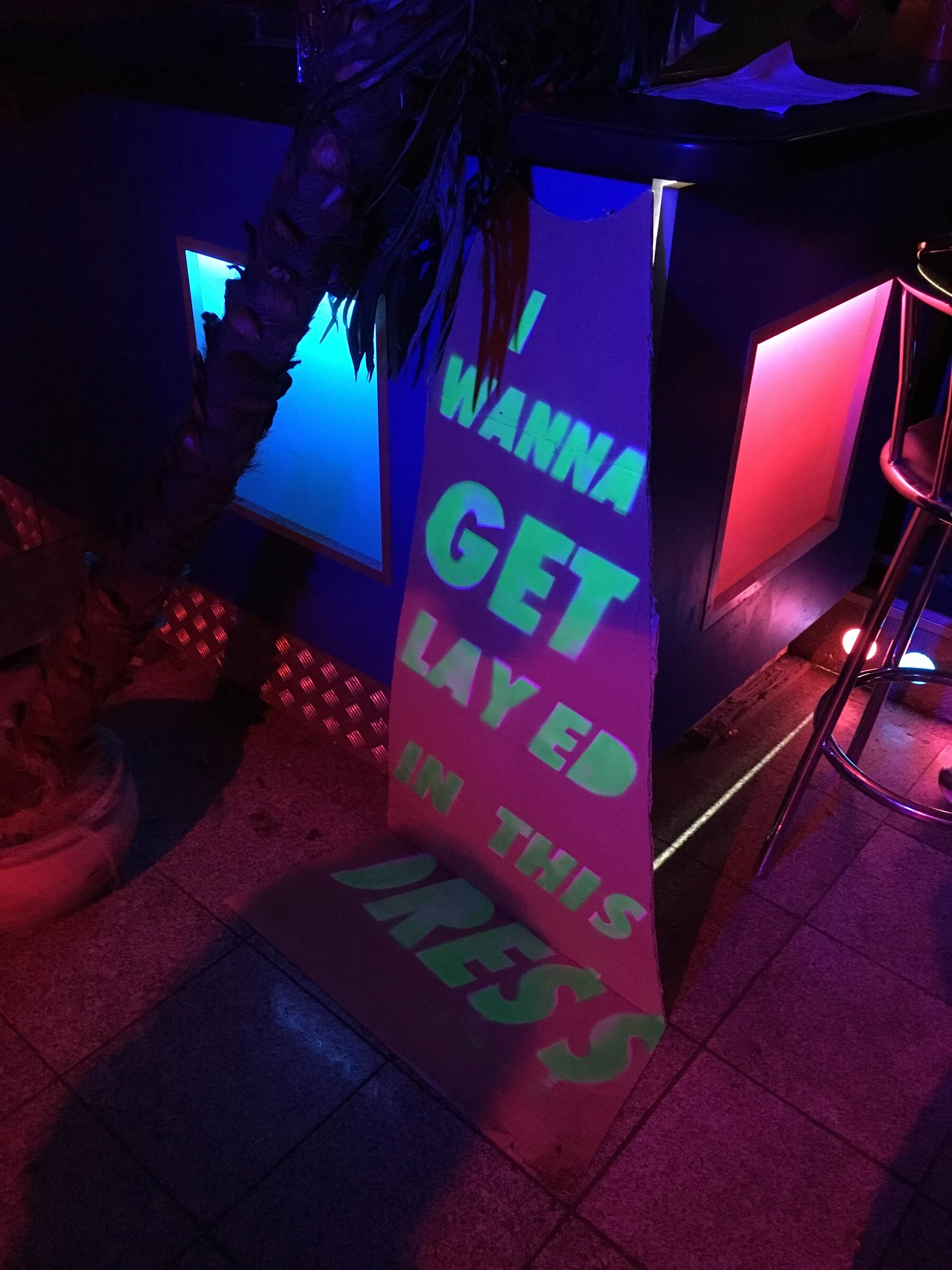 A cardboard dress with green lettering beside a neon-lit bar and a palm tree.