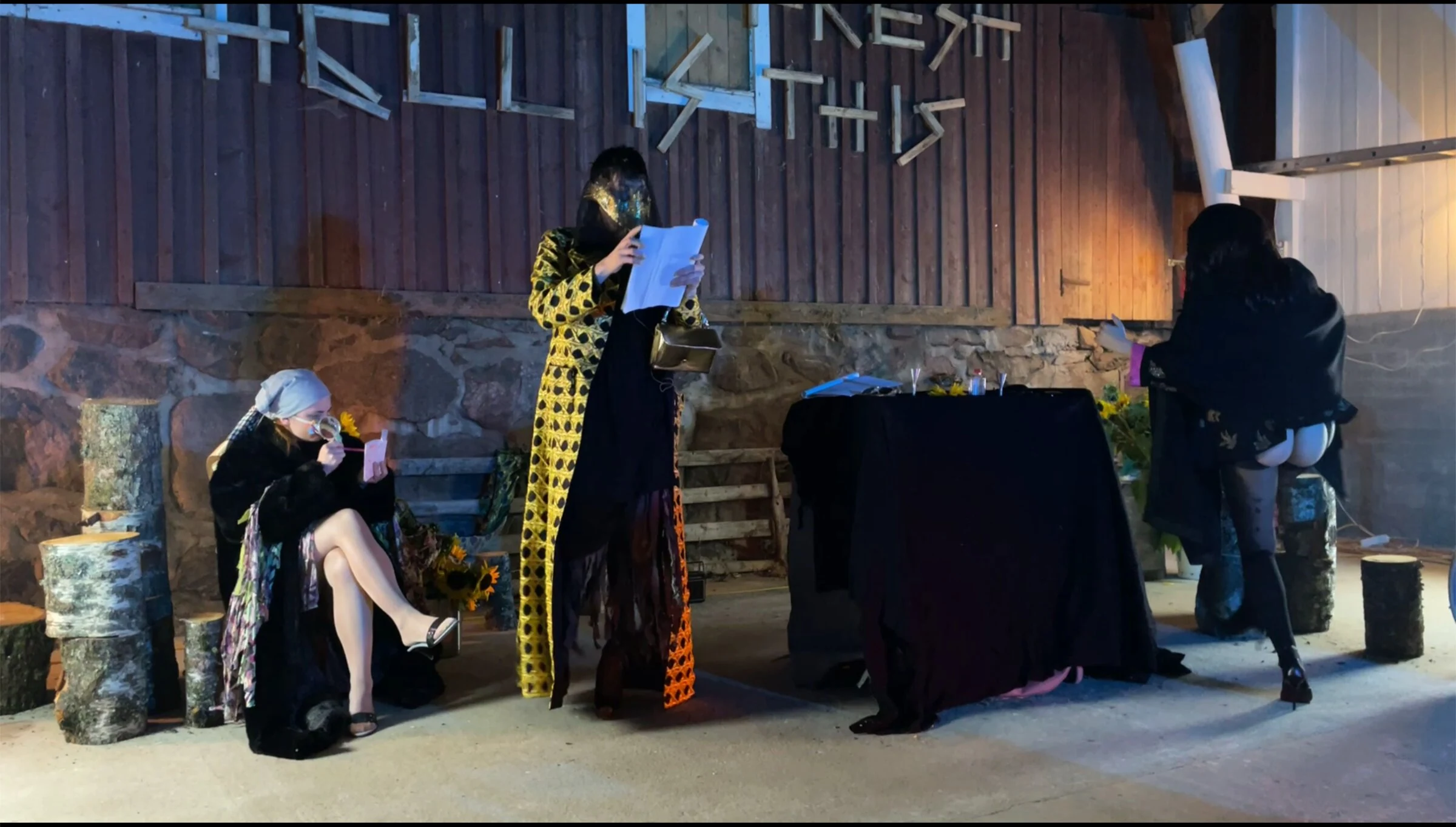 Three masked performers with scripts beside a black-draped table and birch logs.