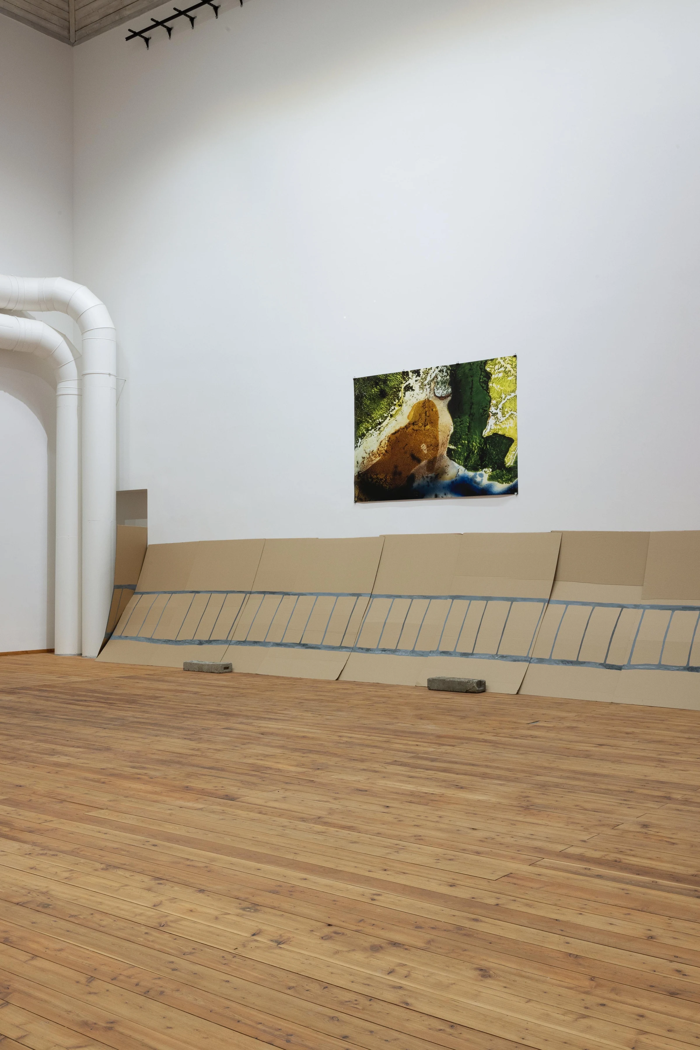 Cardboard fence panels leaning against a gallery wall beneath a painting, beside white ventilation pipes.