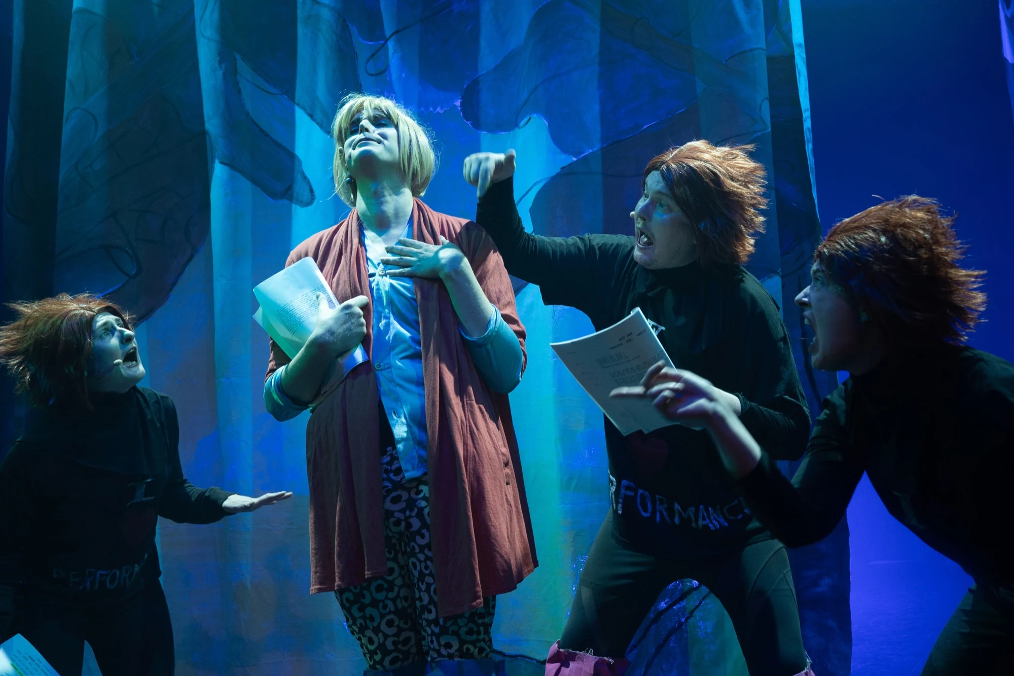 Four performers holding scripts and gesturing under blue stage lighting.