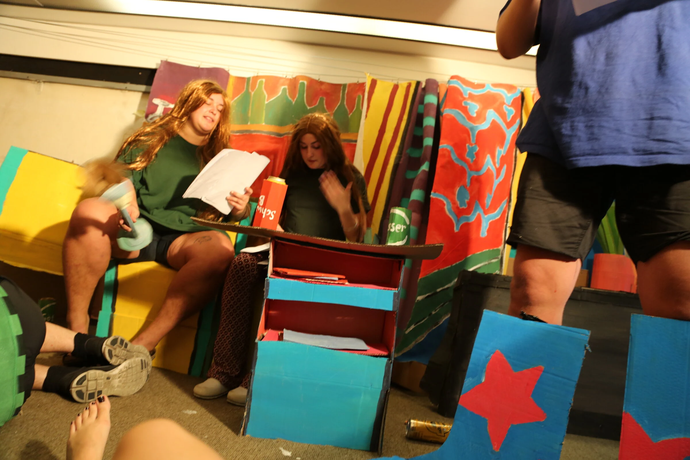 Performers seated on a yellow cardboard sofa behind a handmade table and oversized blue boots.