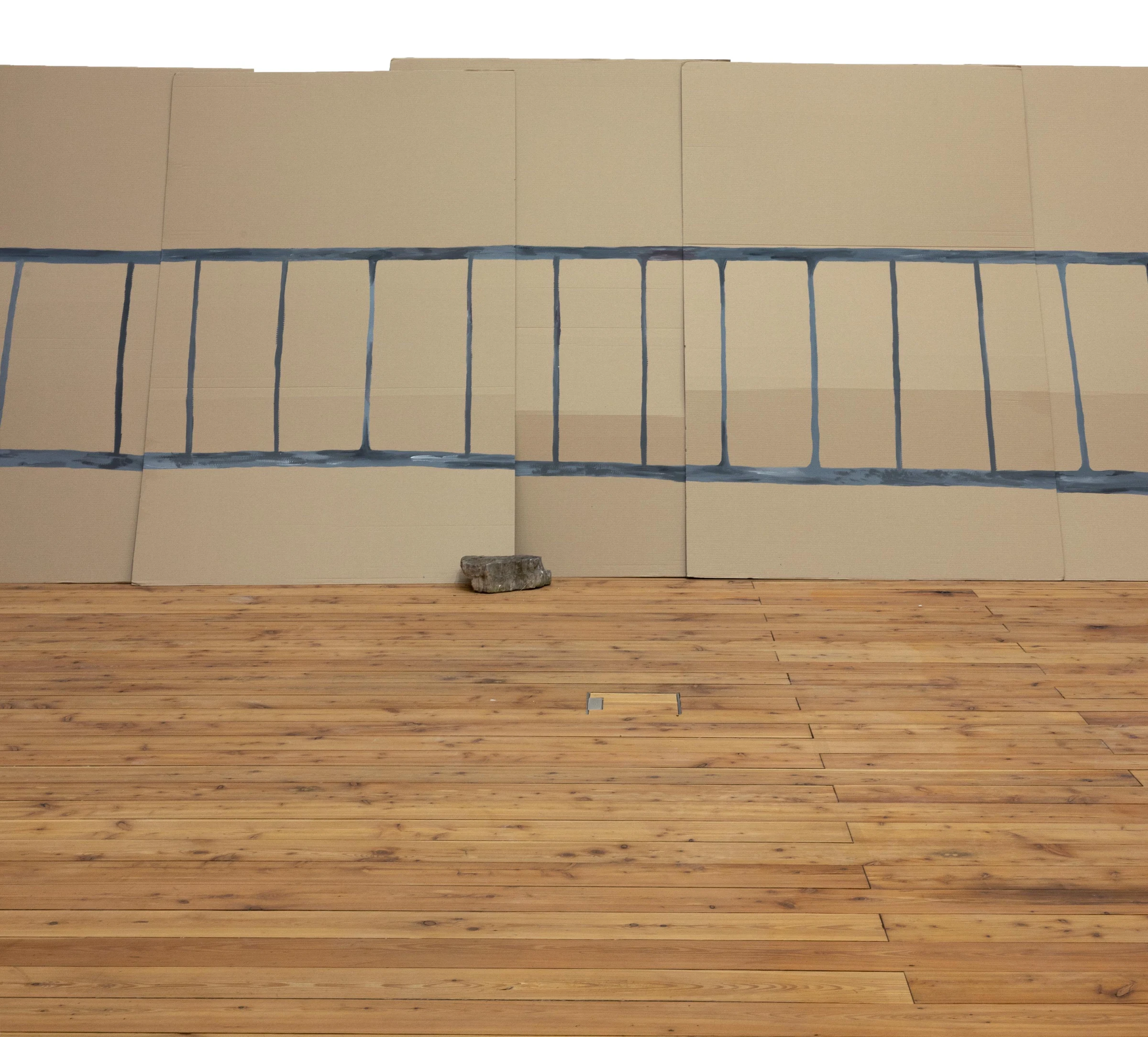 A detail of the blue painted fence across cardboard panels, weighted with a stone on the wooden floor.