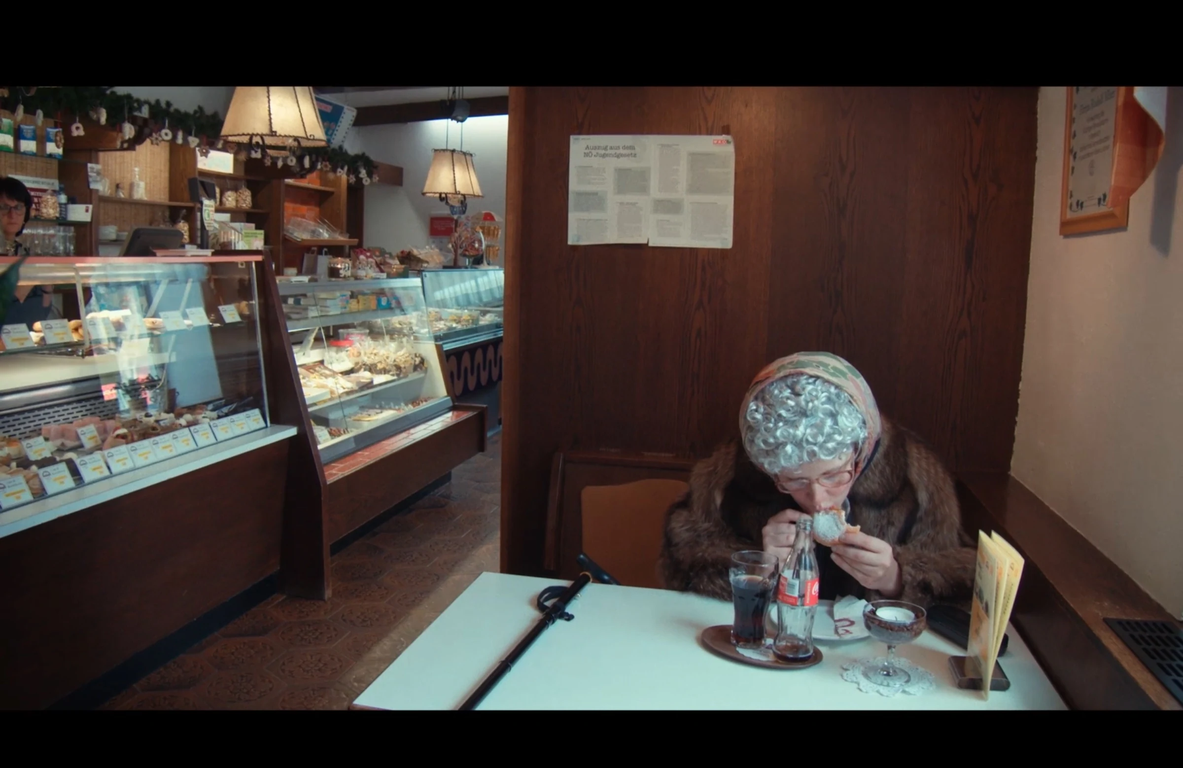 A character wearing a grey wig and headscarf eating a pastry in a café.
