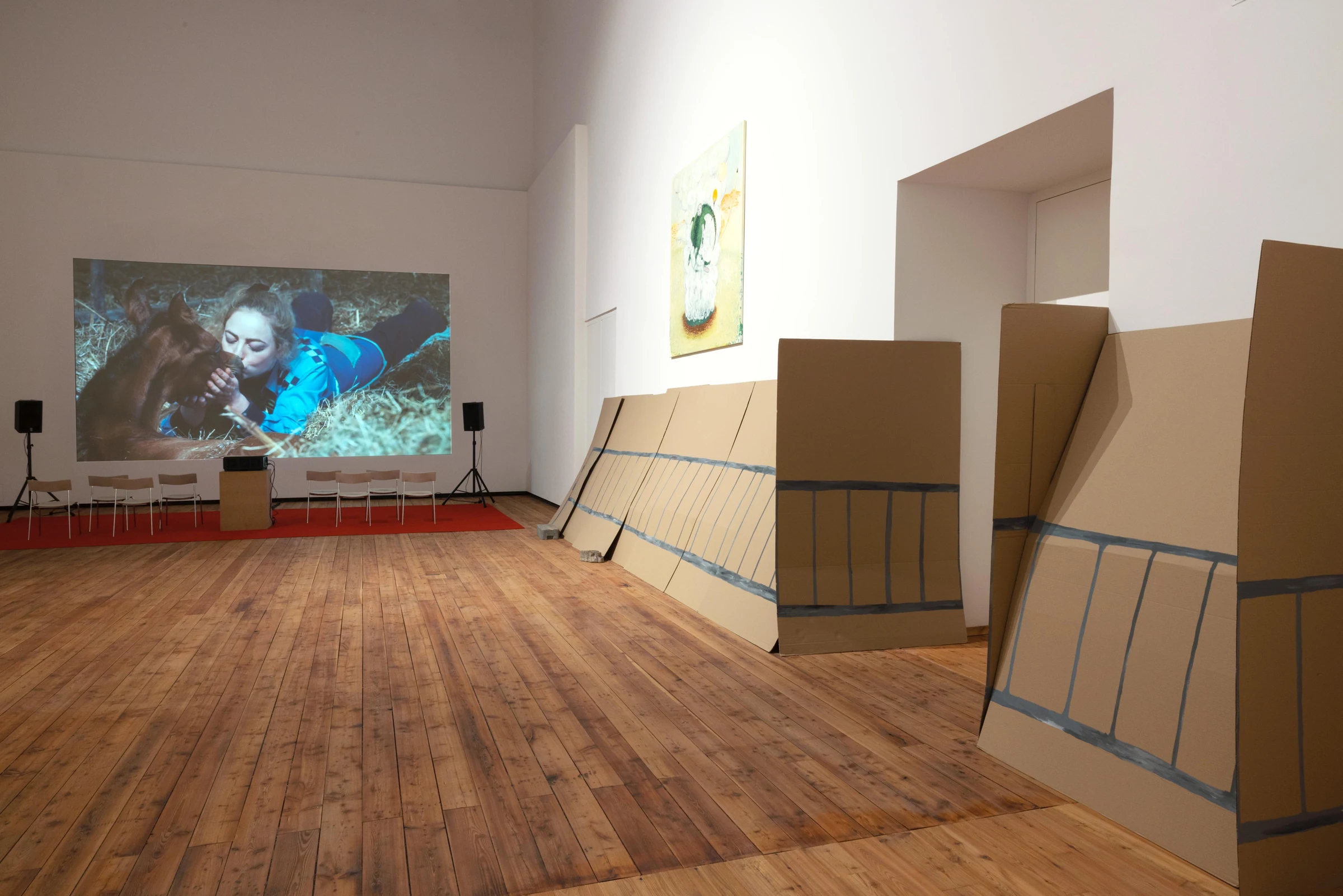 A wide view of the cardboard fence installation with chairs and a projected film at the end of the gallery.