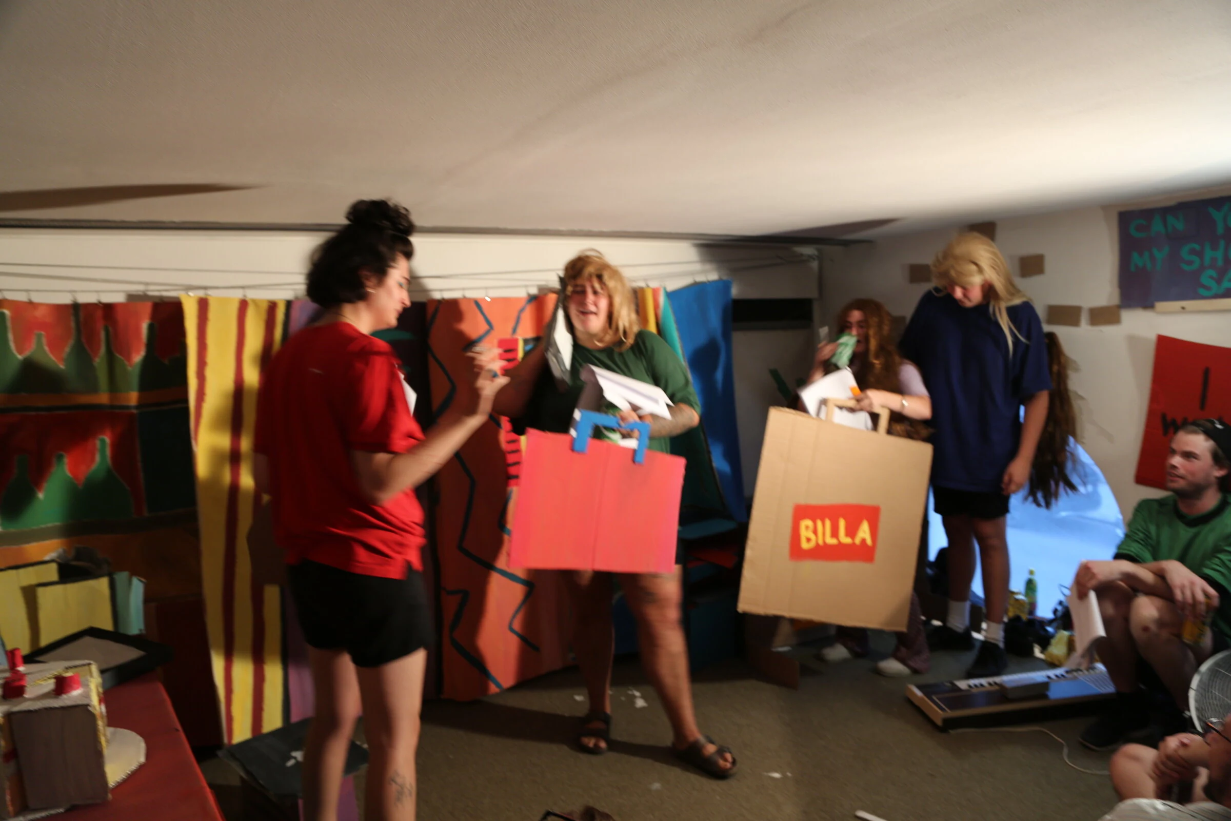 Performers in wigs holding oversized cardboard shopping bags, including one marked BILLA.