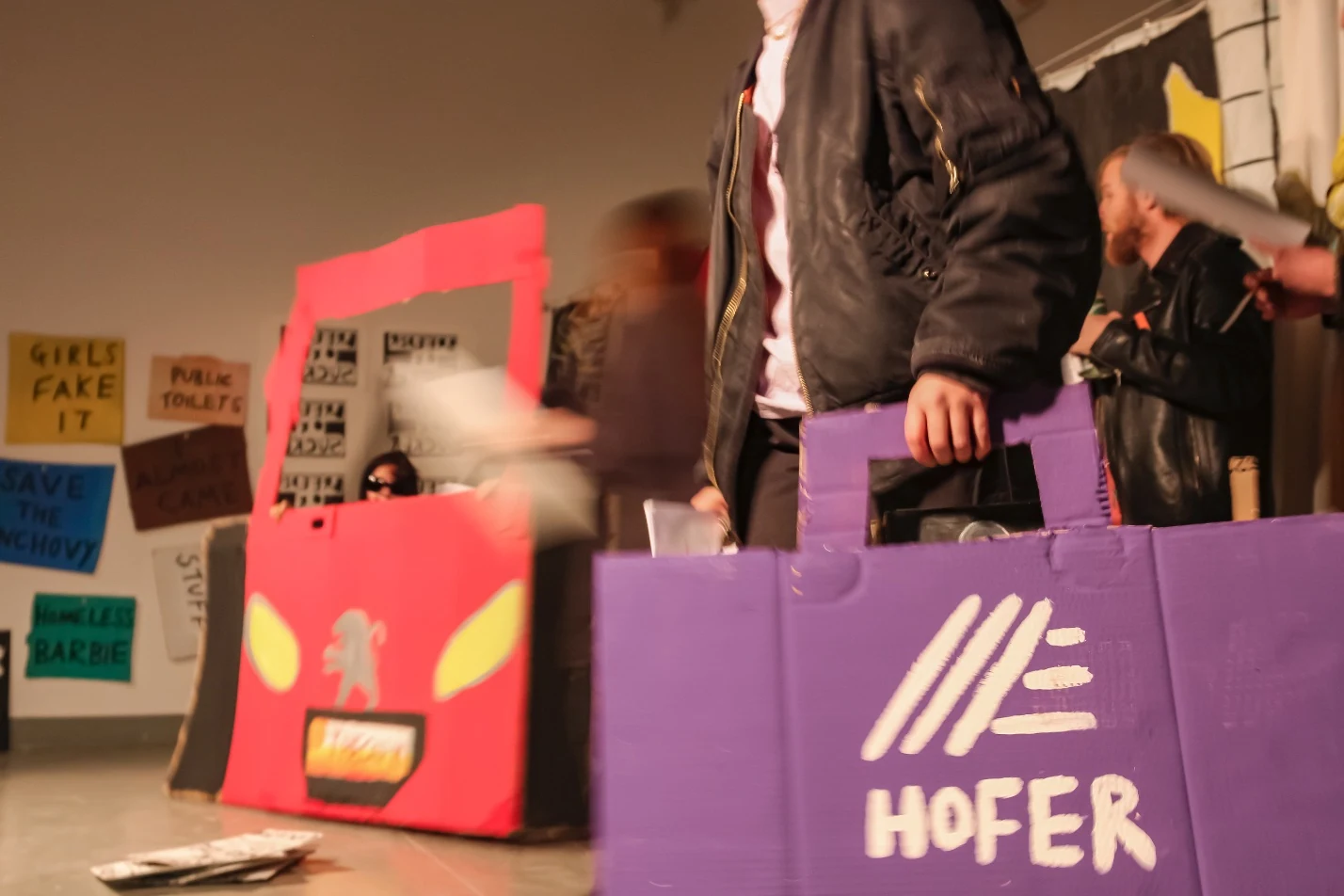 A performer carrying a purple cardboard shopping bag beside a red cardboard car.