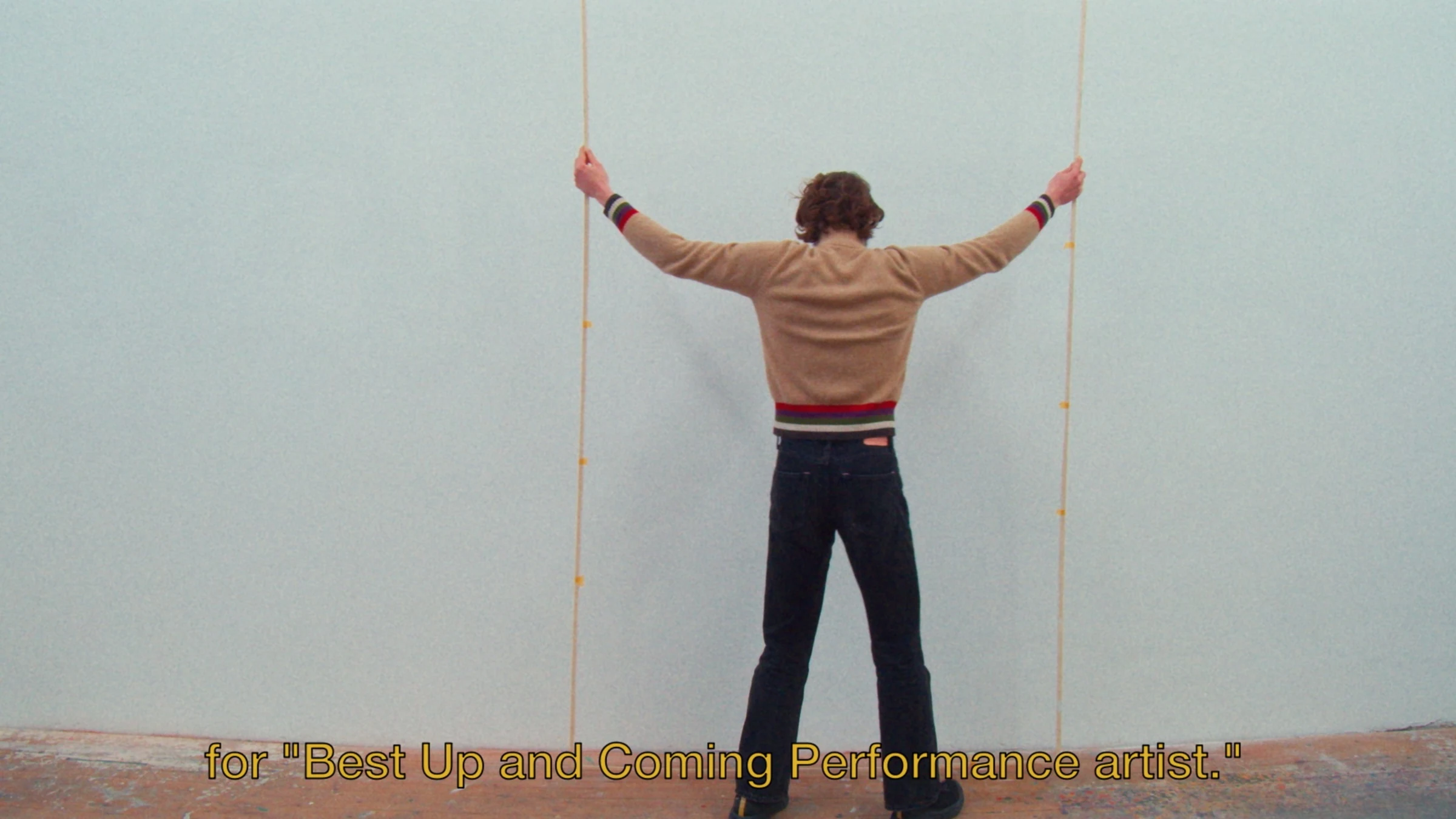 Young Man facing a pale wall with his arms extended, holding two hanging ropes, subtitled Best Up and Coming Performance artist.