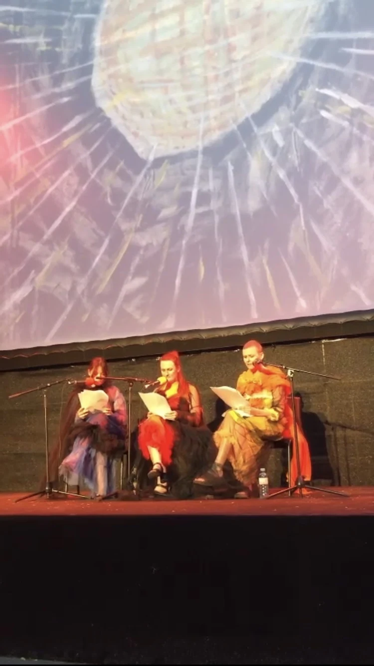 Three seated performers reading beneath a projected disco ball on stage.