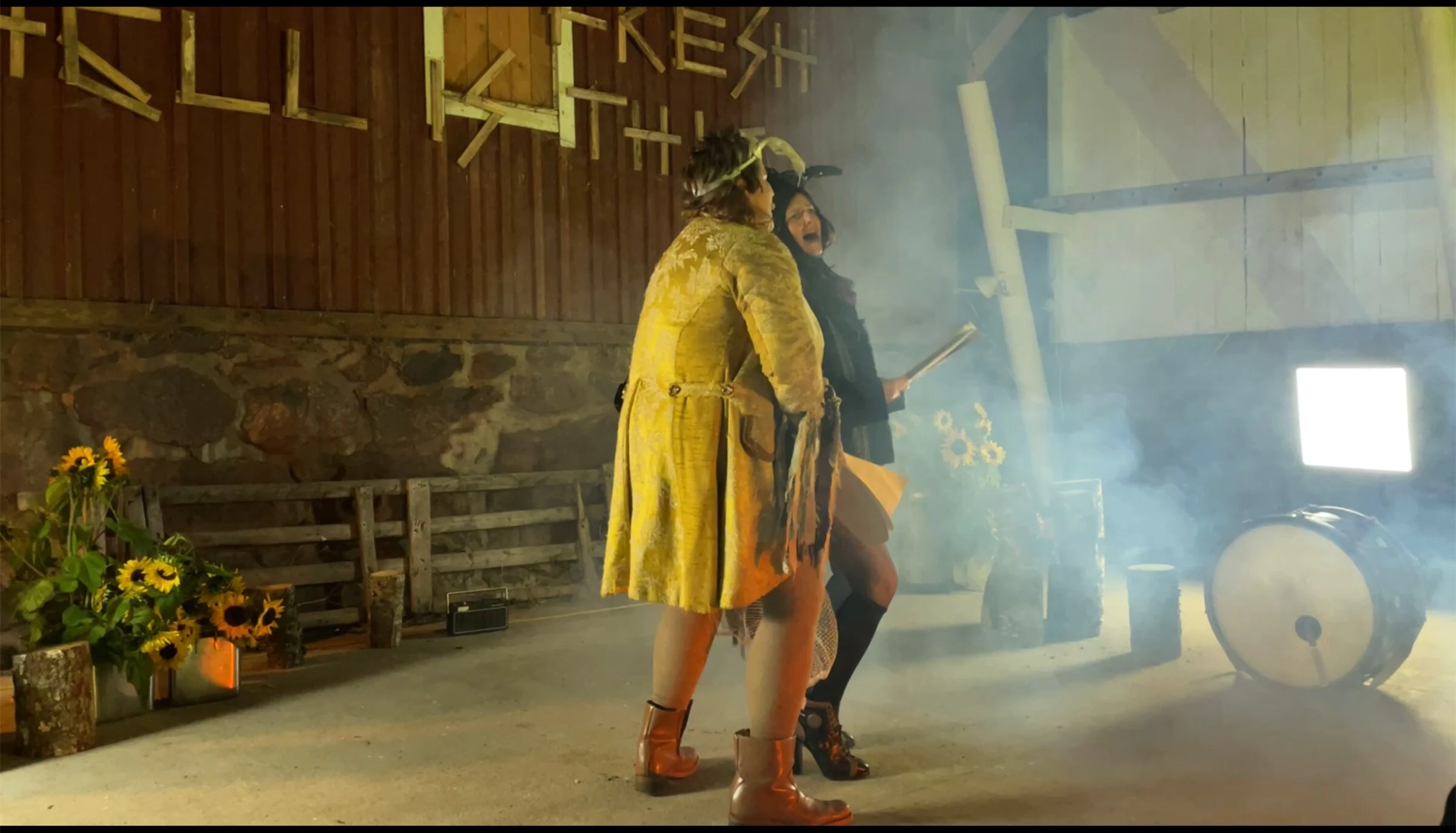 Two masked performers standing together in a barn with sunflowers and a drum.