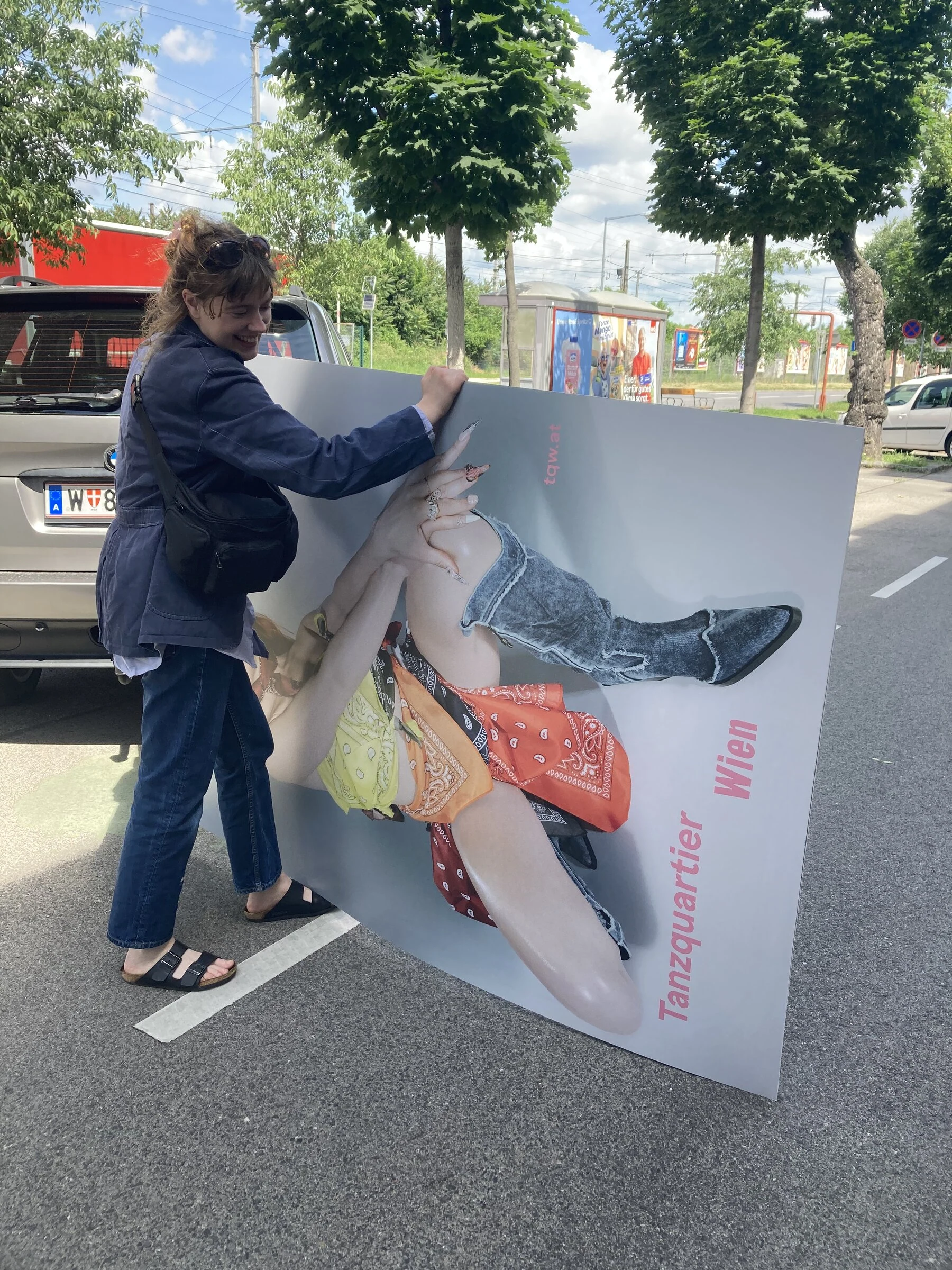 A person holding a large Tanzquartier poster outdoors beside a tree-lined street.