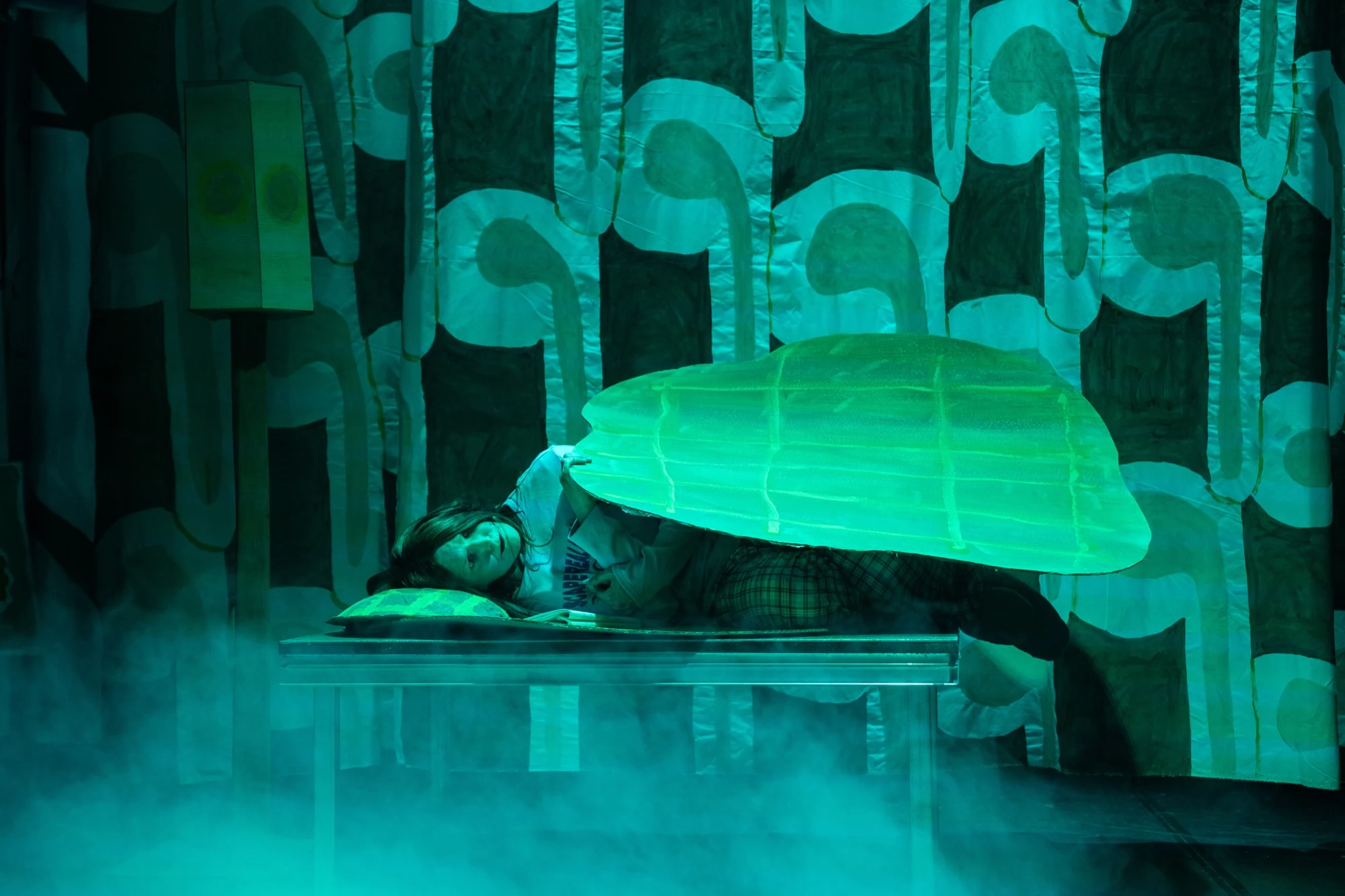 A performer lying beneath an oversized cardboard blanket under green stage lighting.