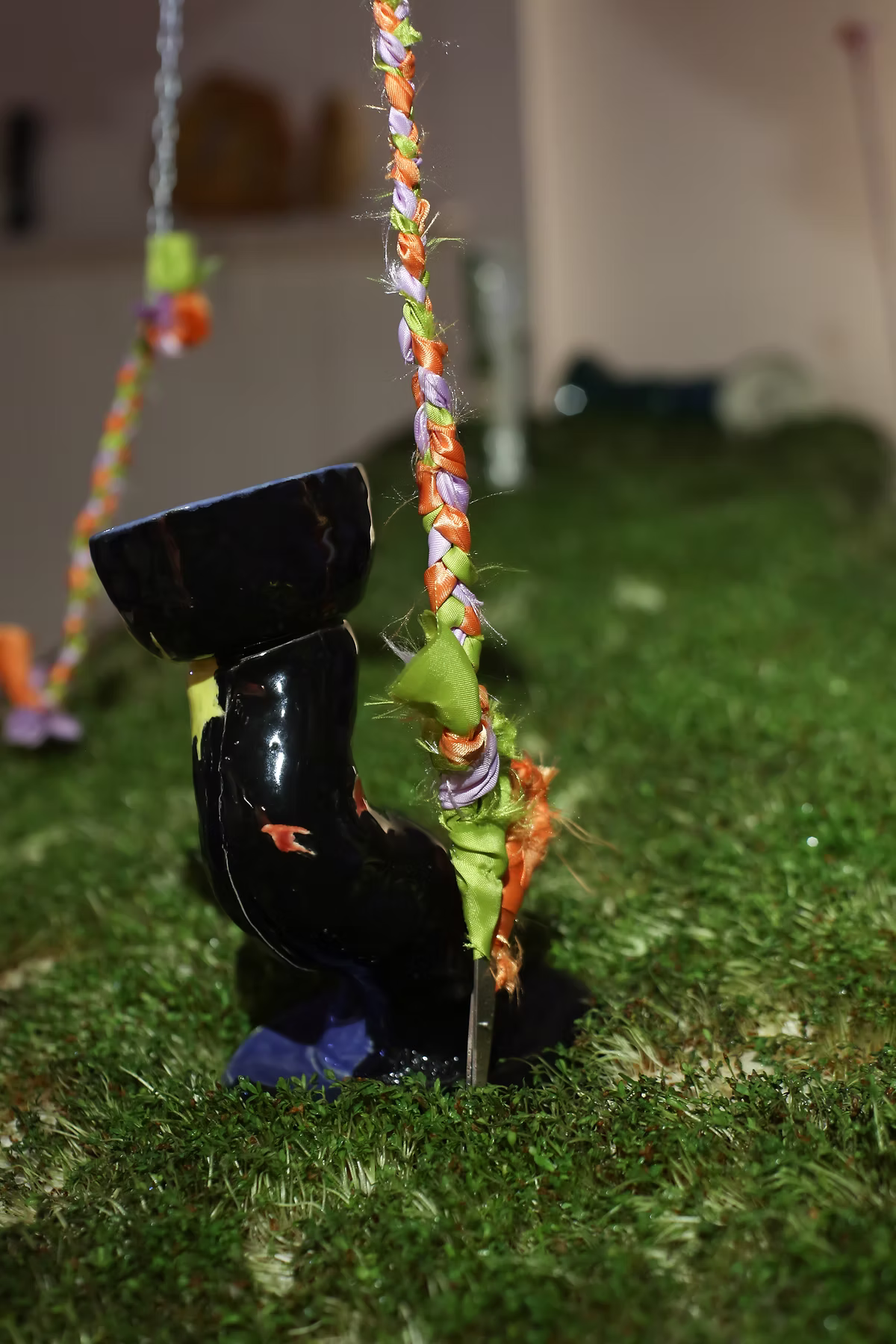 A black ceramic vessel displayed on grass beneath colourful hanging ropes.