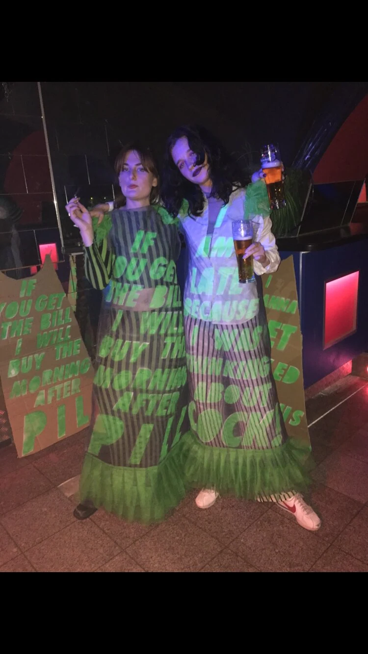 Two performers wearing dresses with projected green lettering under blue and purple light.