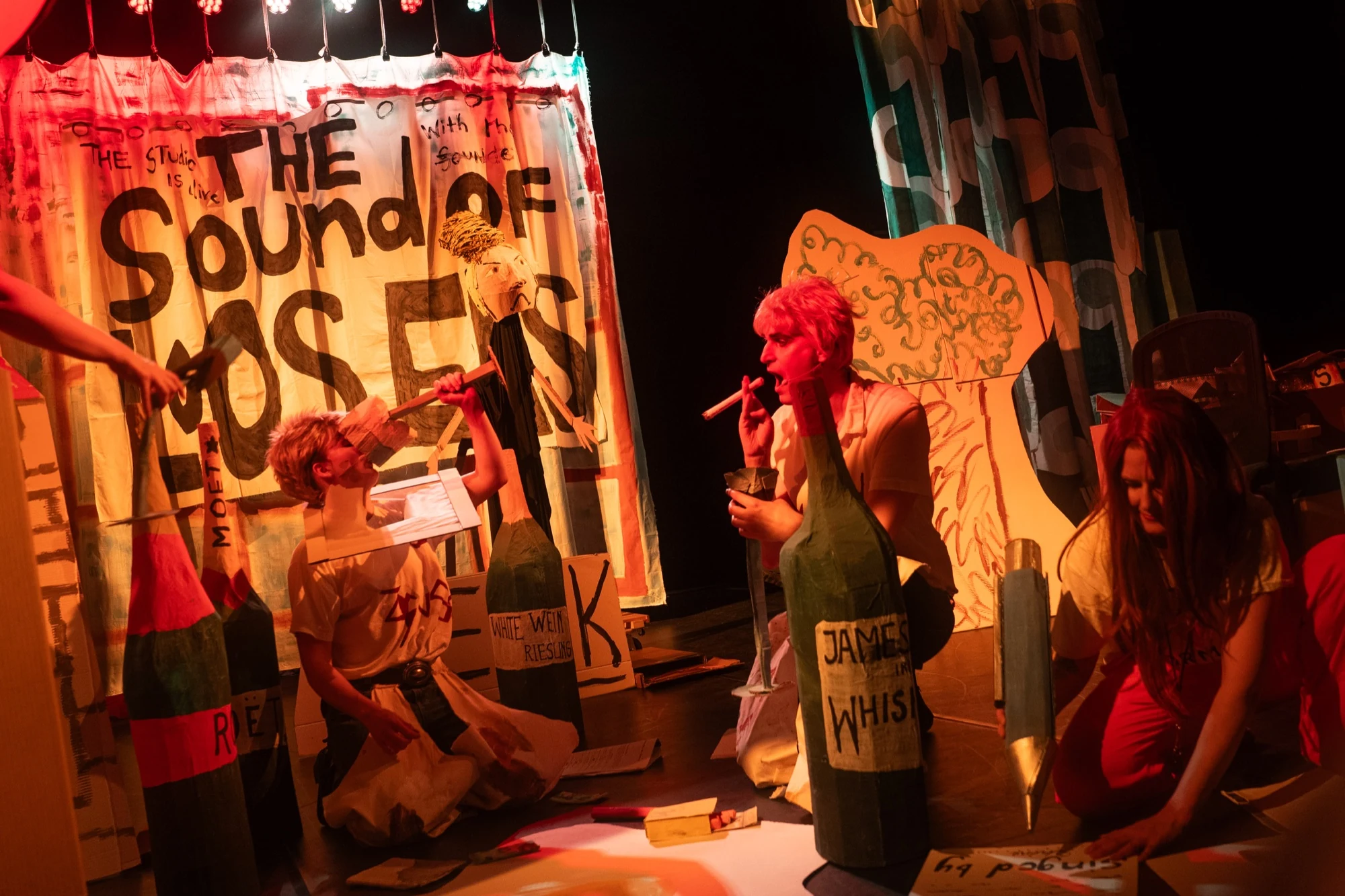 Performers among oversized cardboard bottles and scenery under red stage lighting.