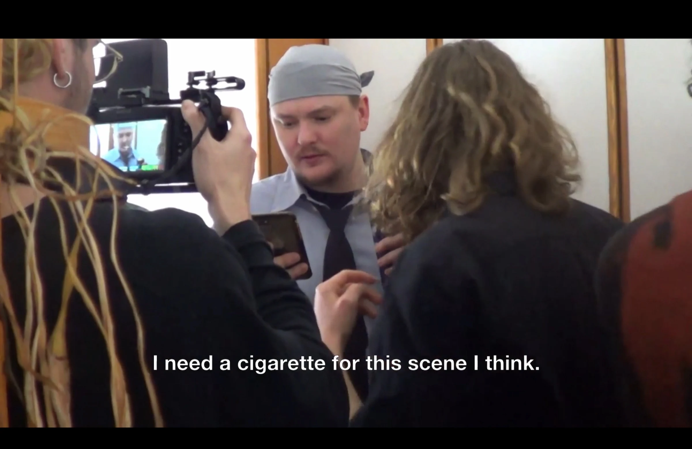 A camera operator films a performer in a grey headscarf and tie as the crew prepares a scene.