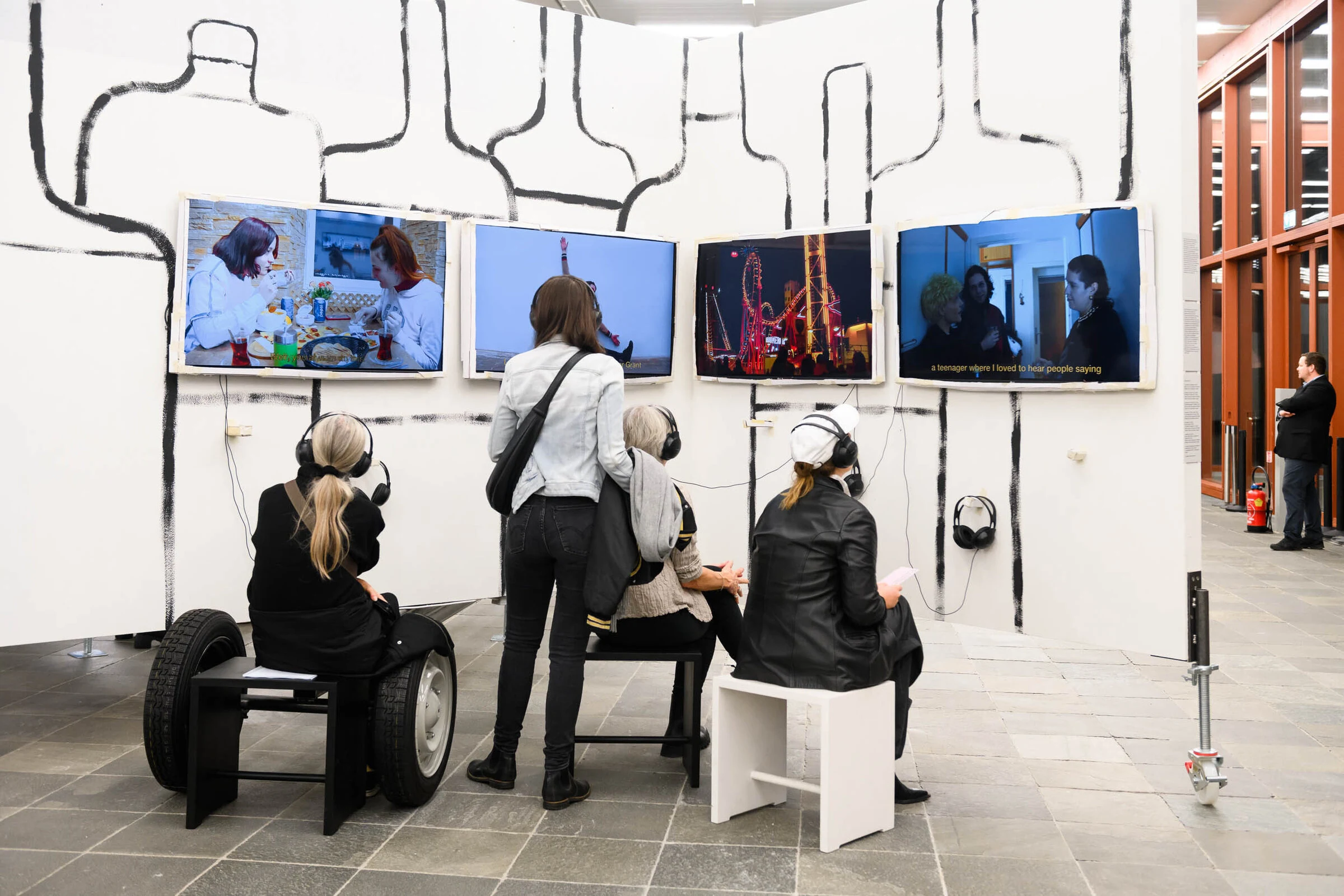 Visitors wearing headphones in front of four LOCATION video screens mounted on a wall painted with bottle outlines.