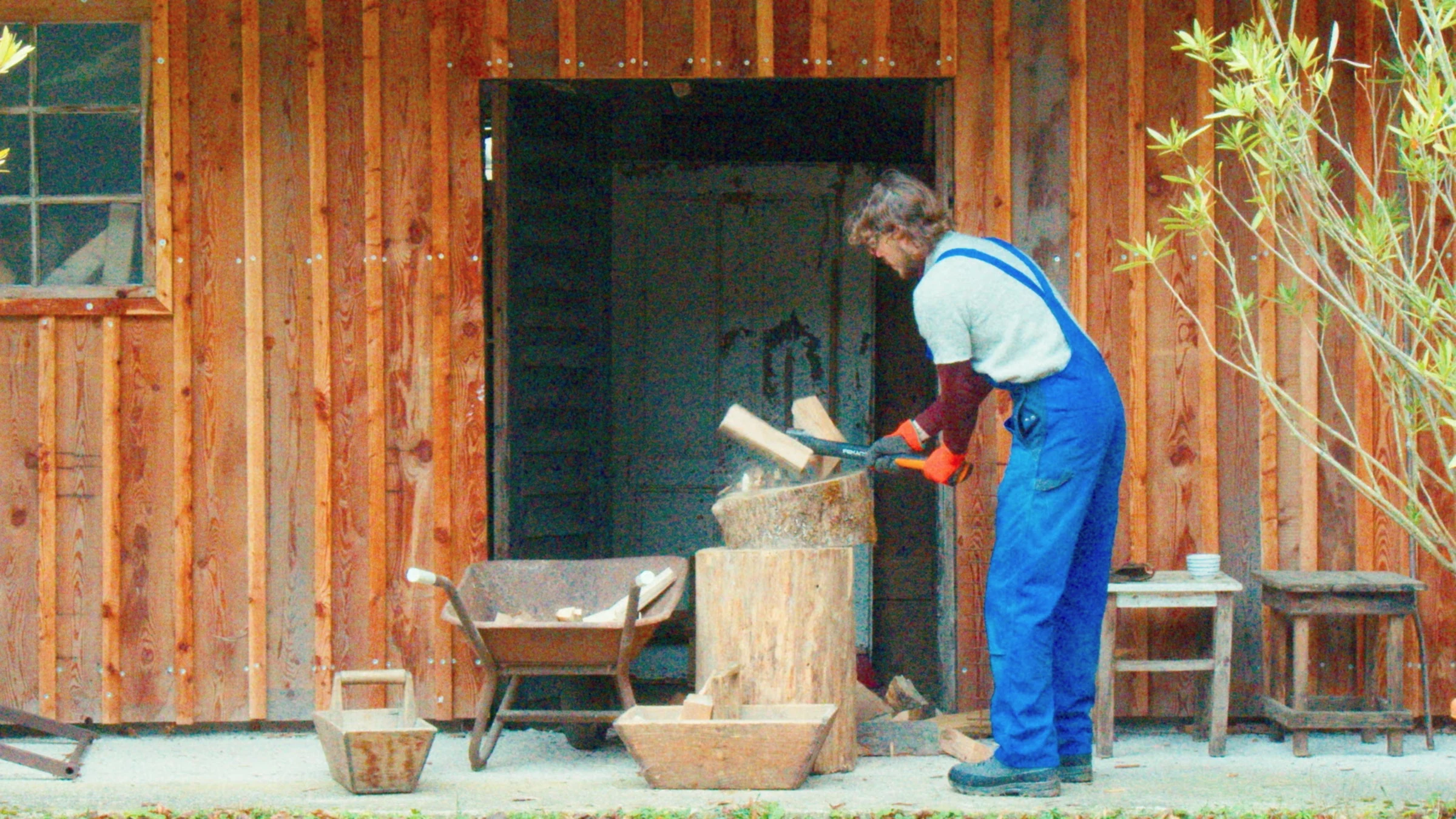 A performer in blue overalls chopping wood outside a wooden shed.