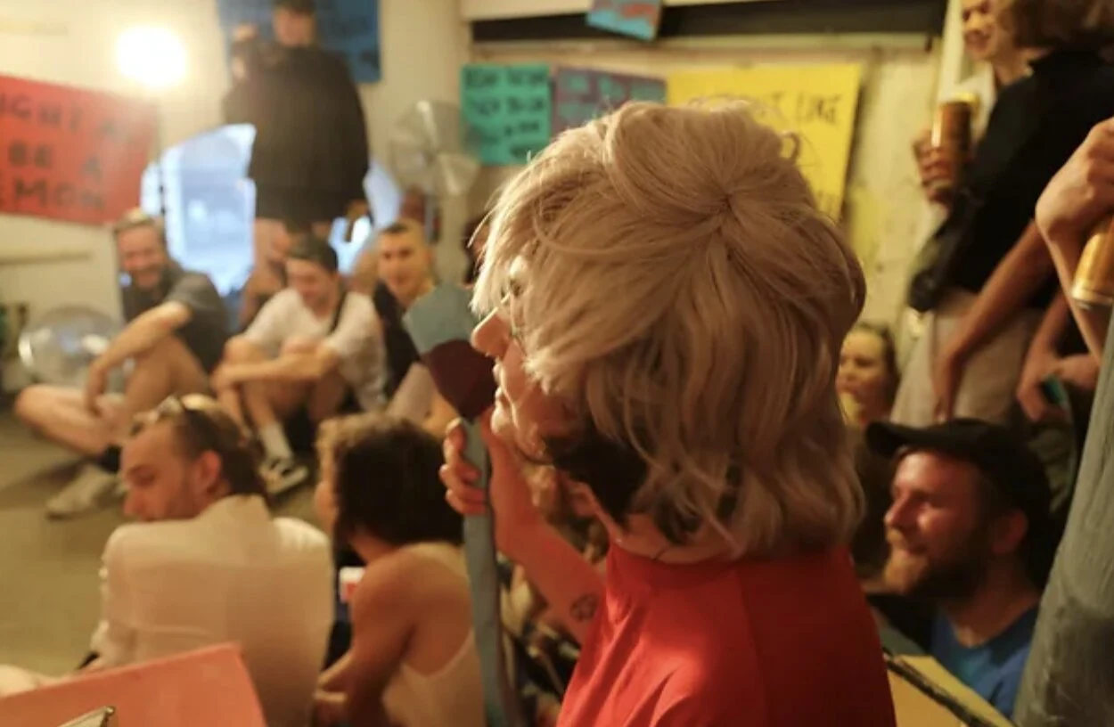 A performer in a blonde wig and red shirt surrounded by the seated audience.