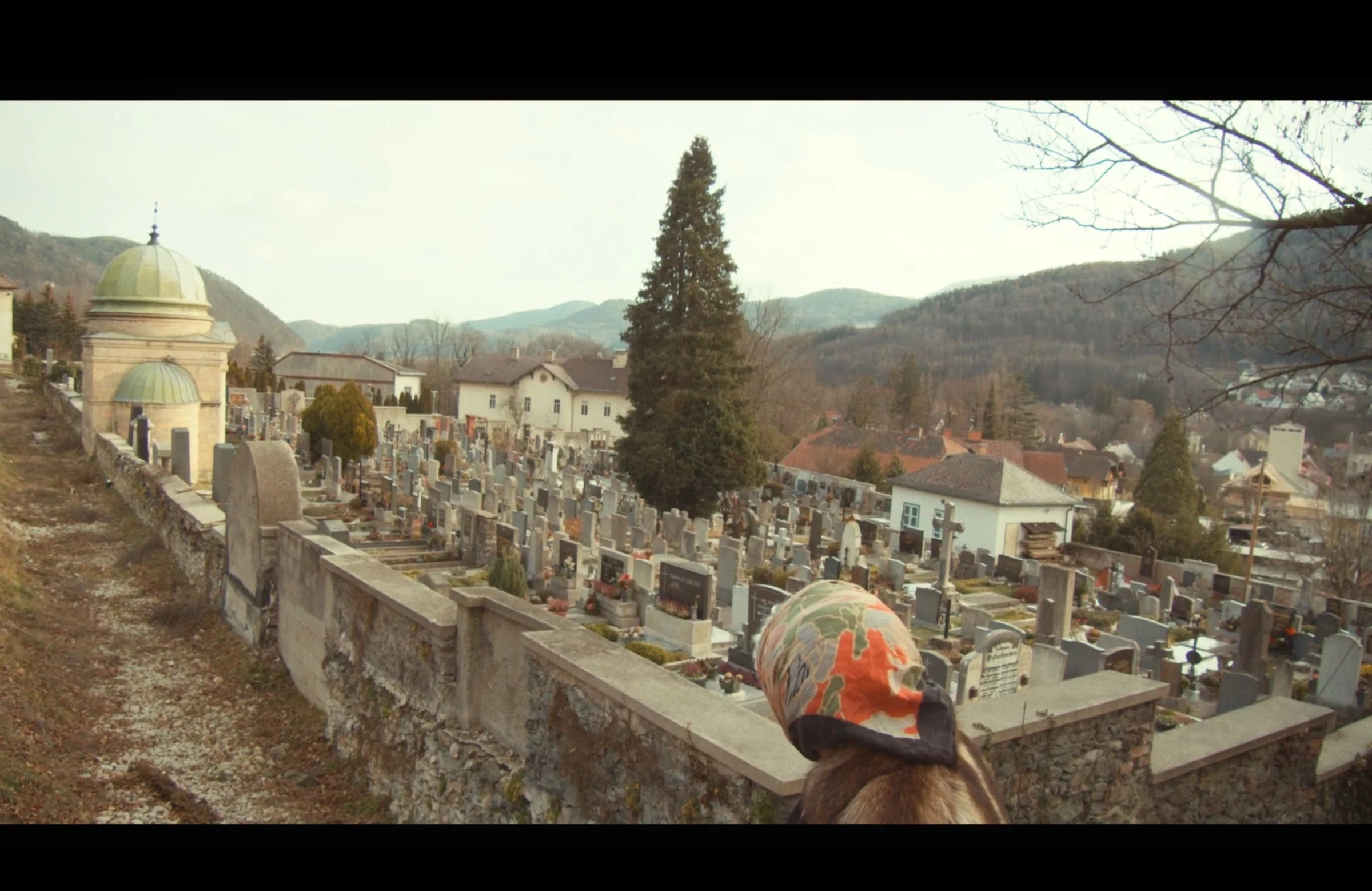 A character in a headscarf overlooking a cemetery and a wooded valley.
