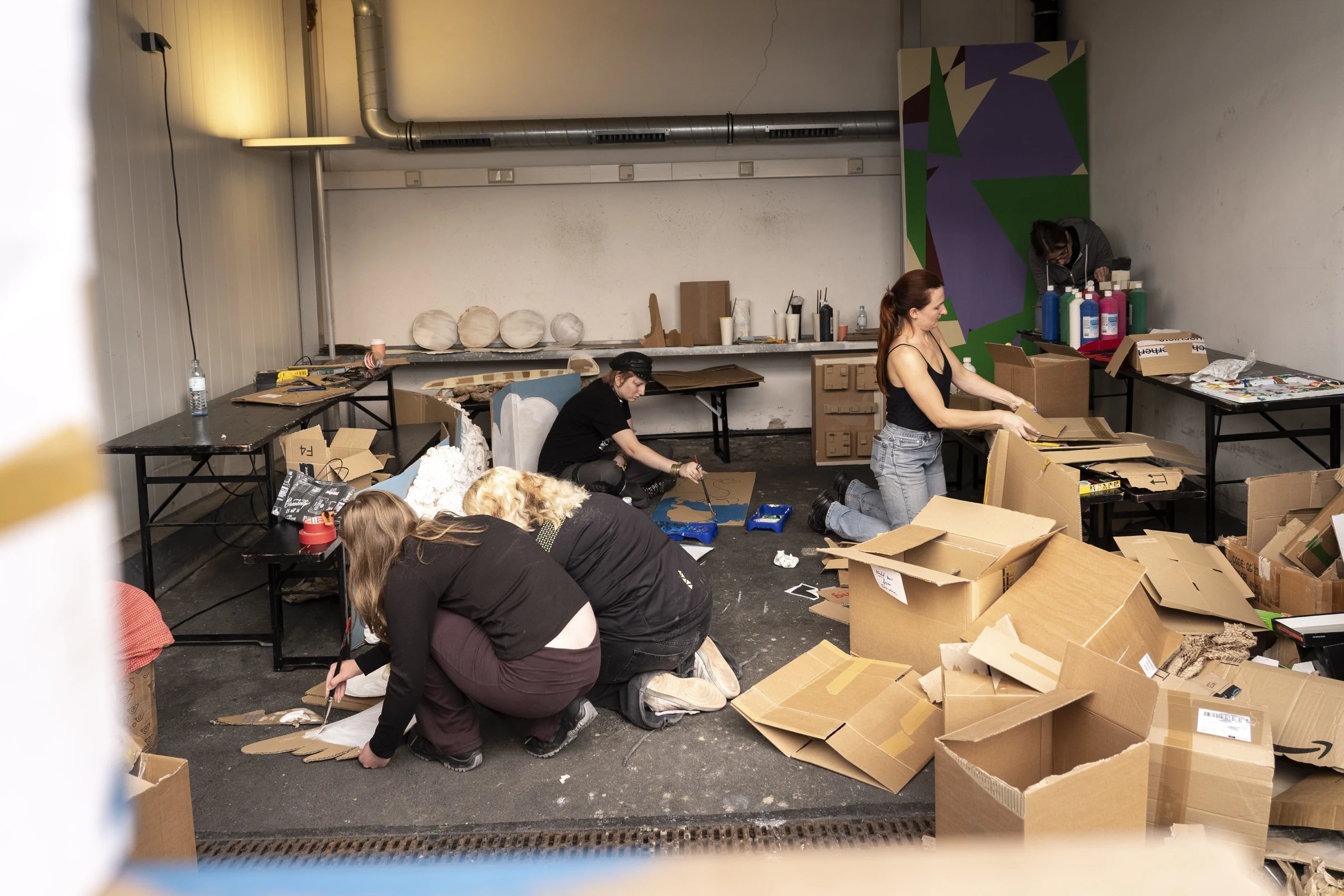 Workshop participants painting and assembling cardboard props among boxes and art materials in a studio.