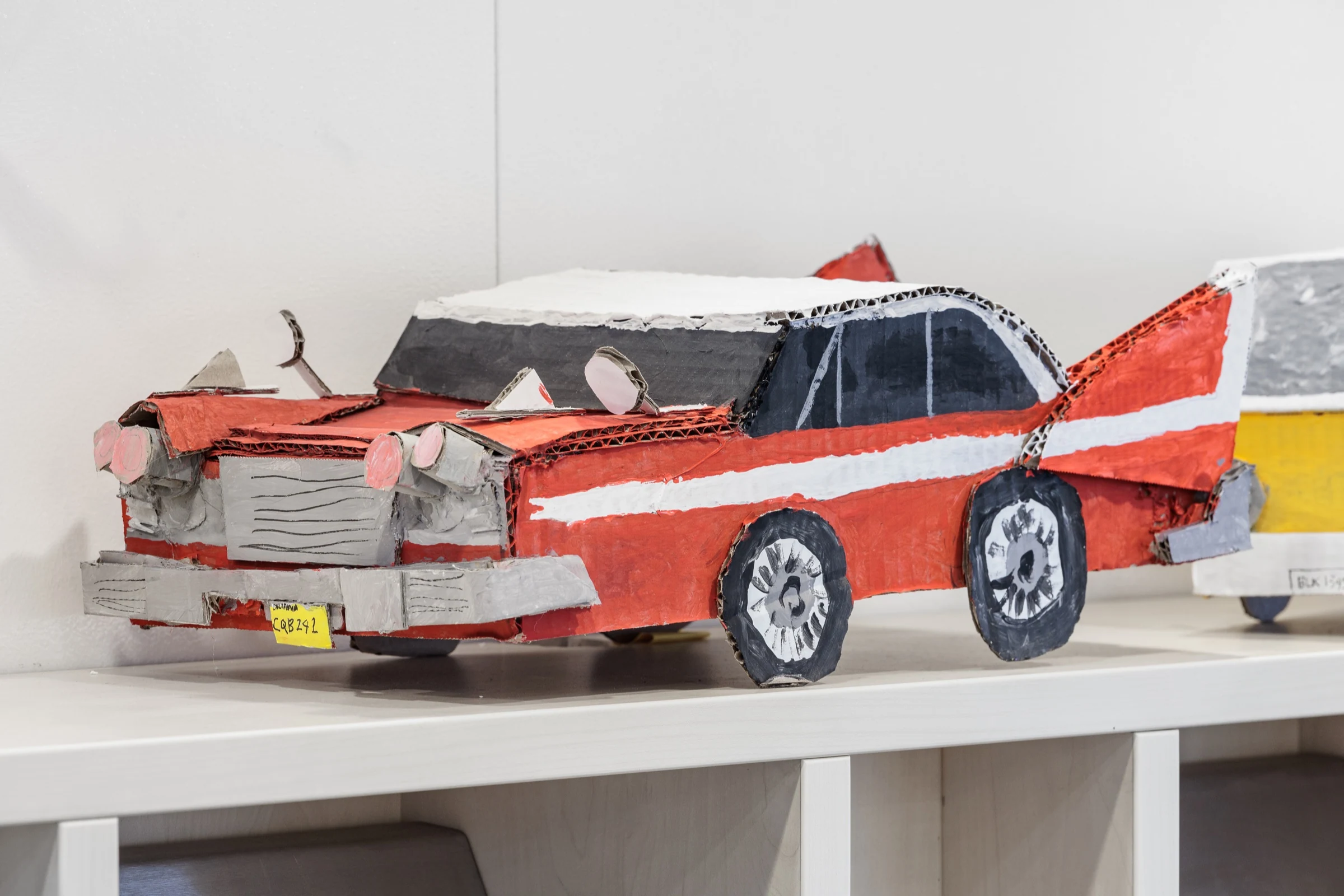 A red and white handmade cardboard car model on a shelf.