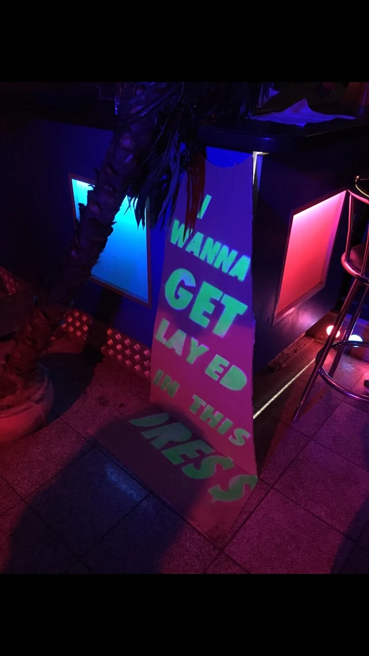 A cardboard dress with green lettering displayed beside a neon-lit bar.