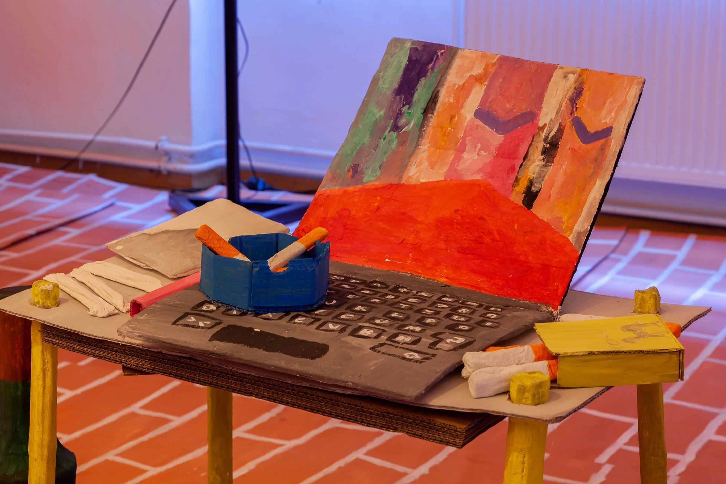 A detail of the painted cardboard laptop and table with an ashtray and yellow objects.