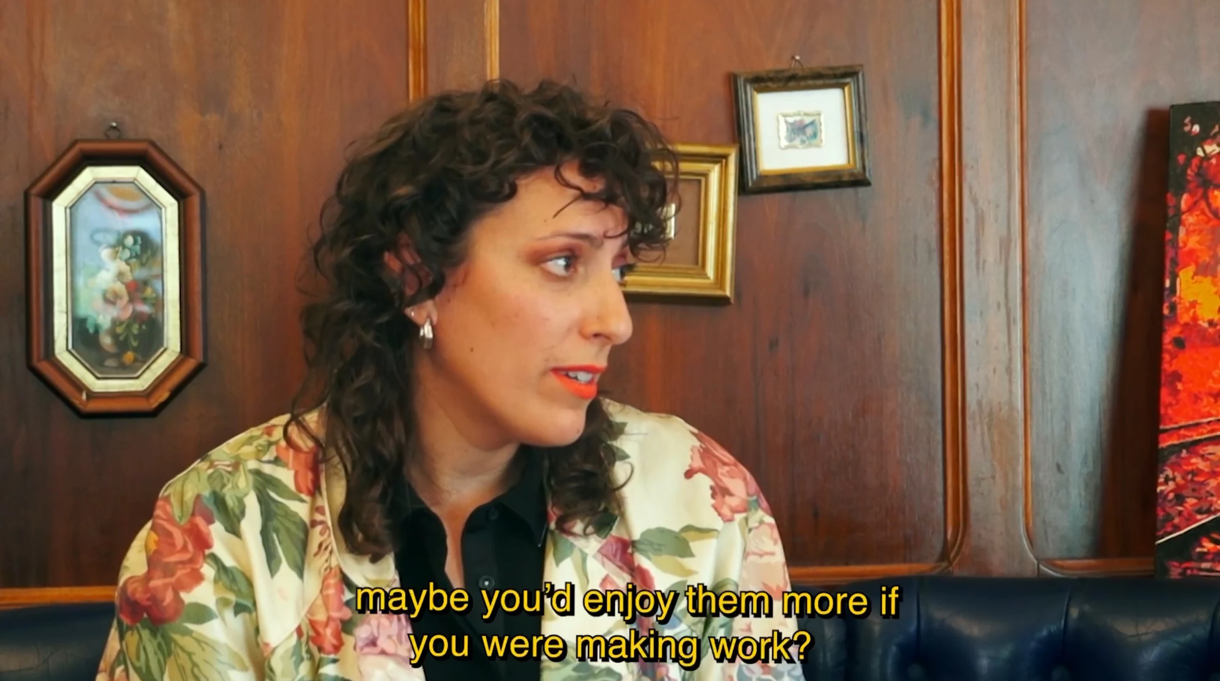 A curly-haired character wearing a floral jacket in a wood-panelled café, asking about making work.