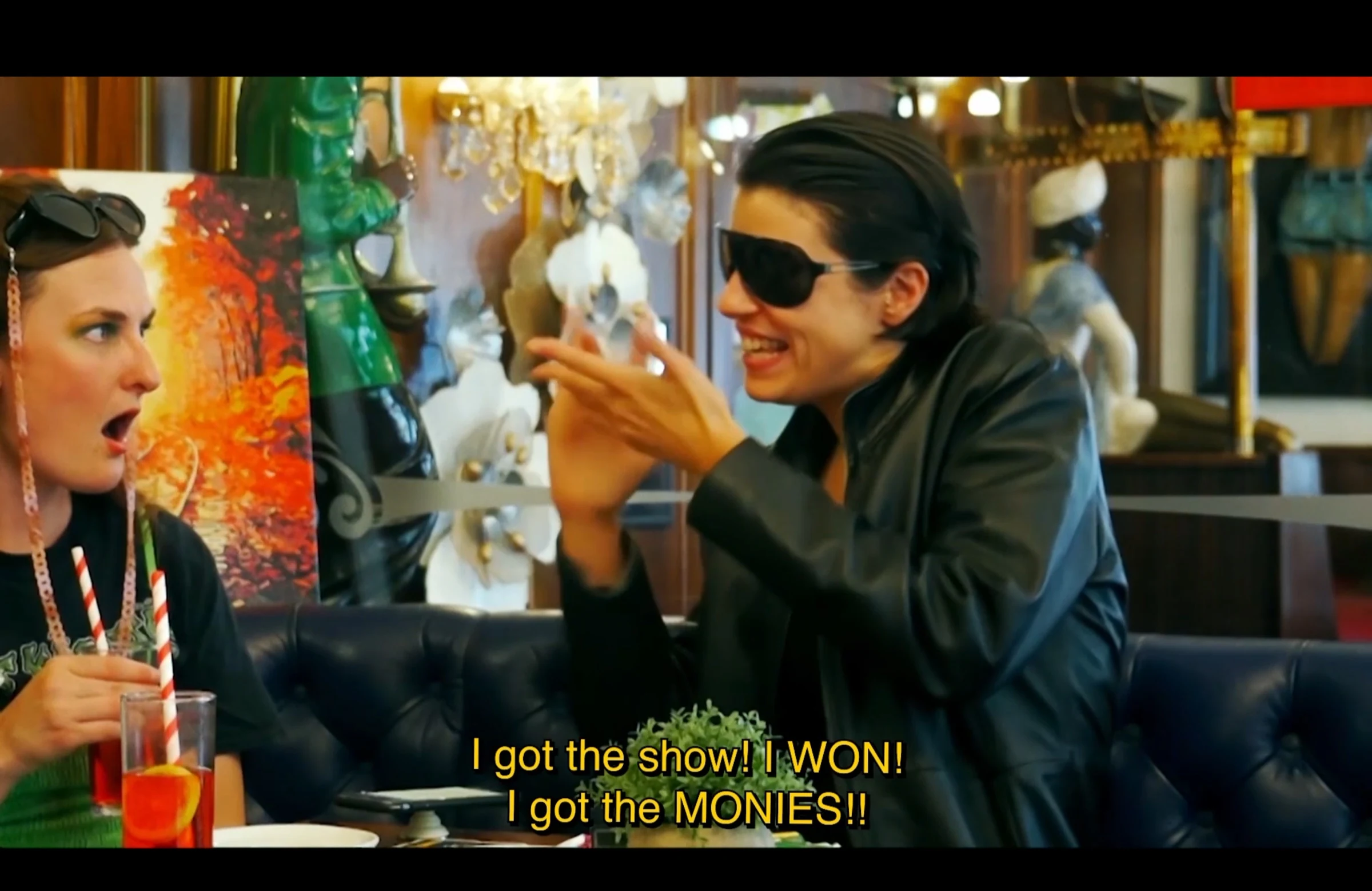 Two characters talking in a café, one celebrating winning a show.