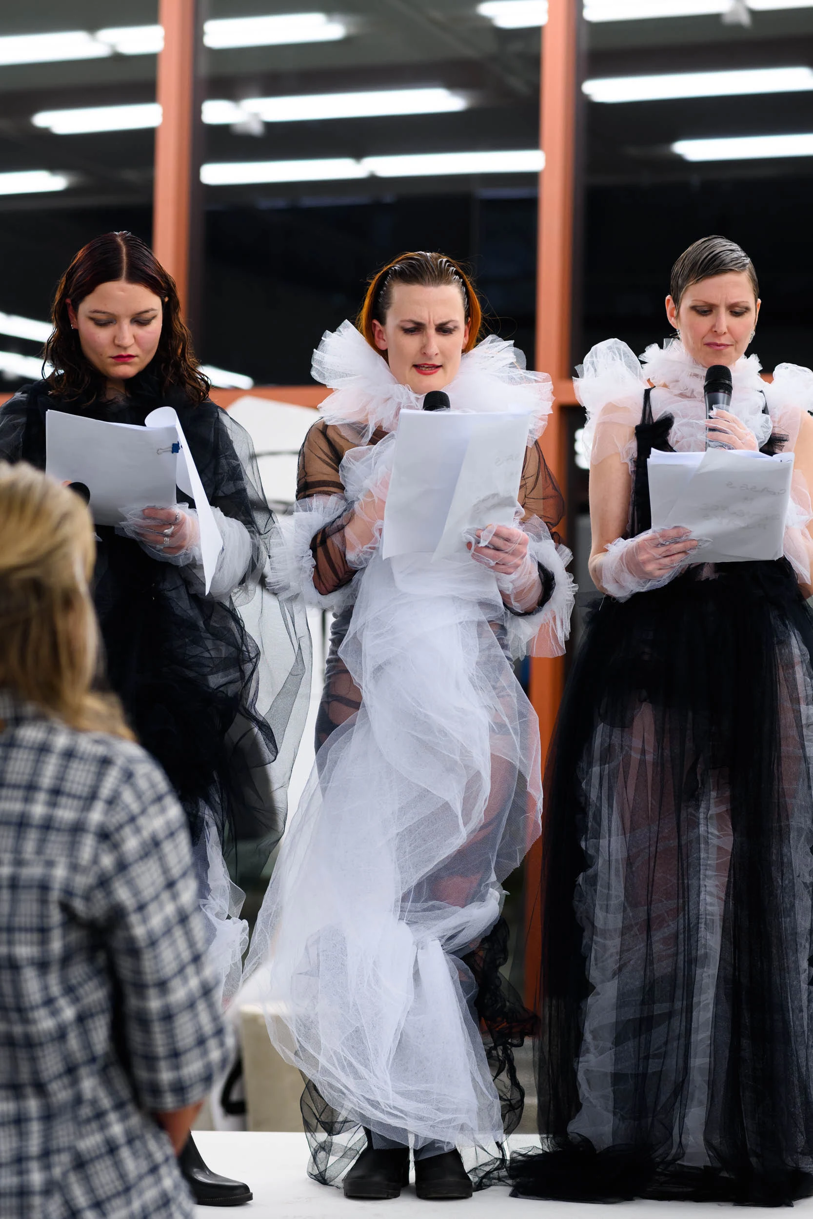 Three performers in black and white tulle costumes reading scripts into microphones in a gallery.