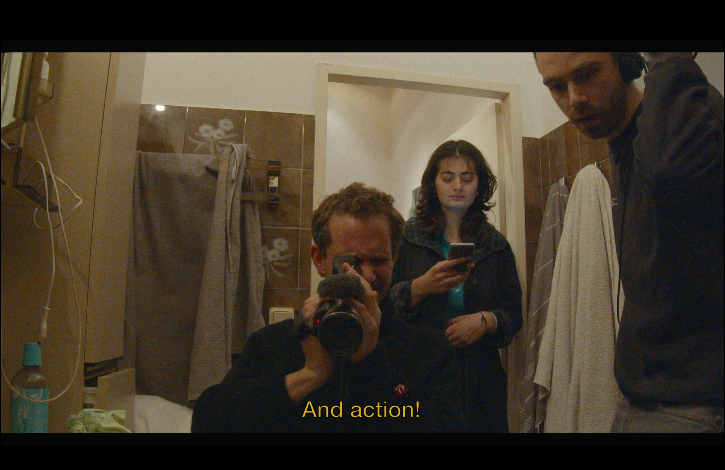 A camera operator filming in a tiled bathroom with two people behind him, subtitled And action!