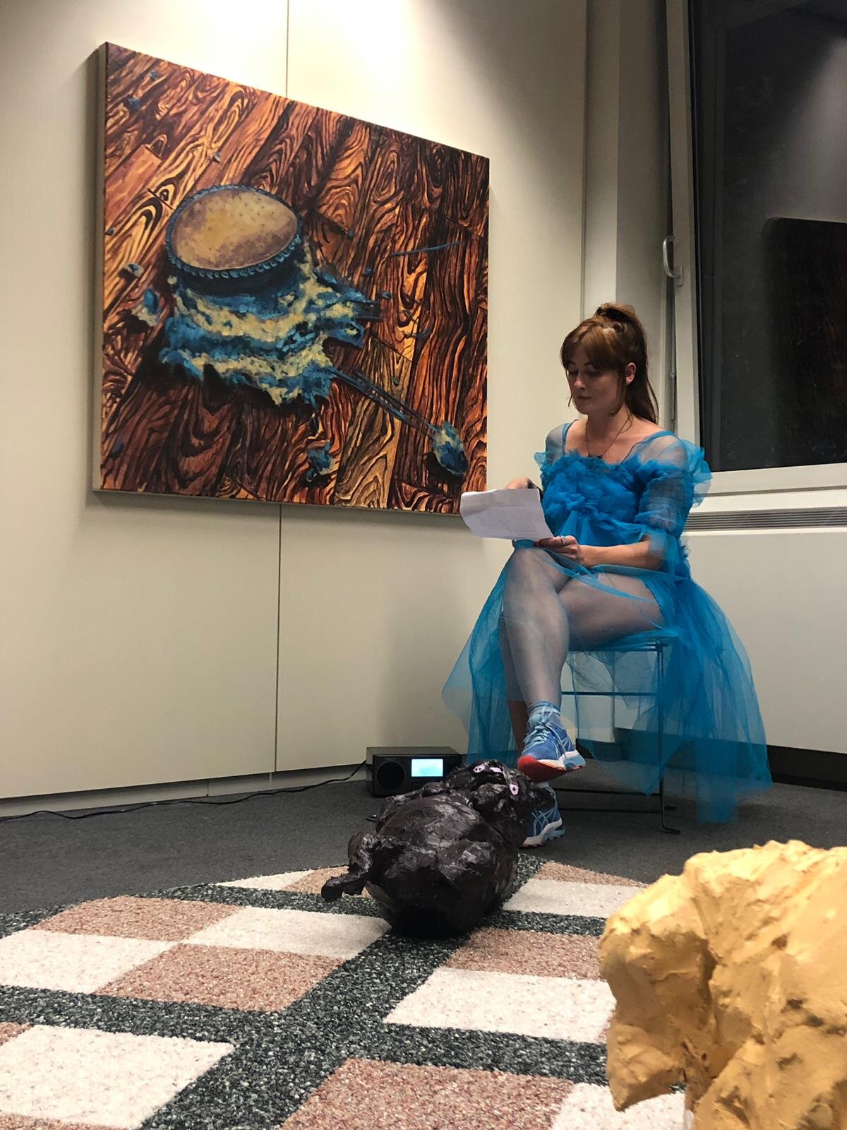A performer seated in a blue tulle costume with sculptures in the foreground.