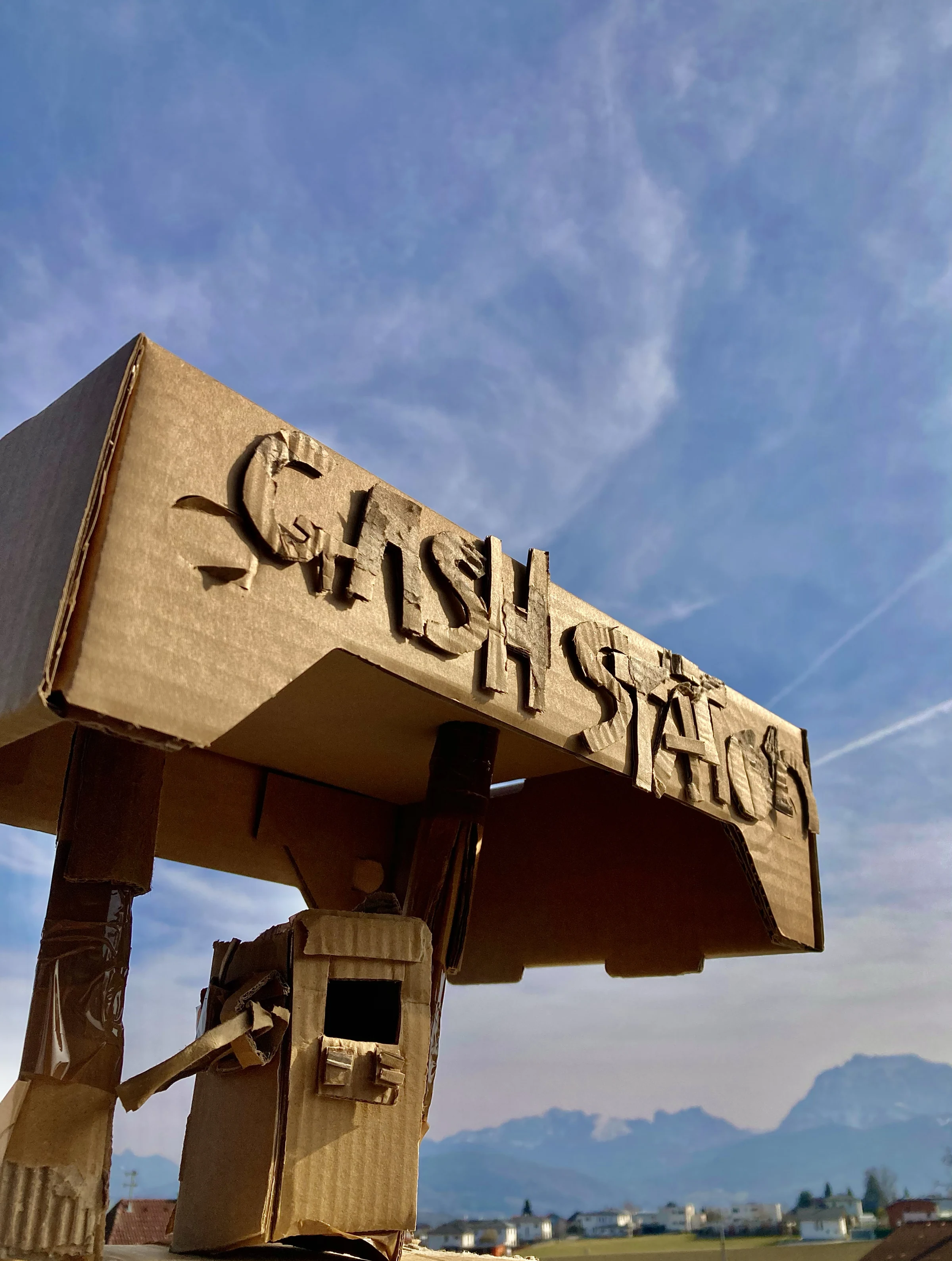 A cardboard GASH STATION model photographed outdoors beneath a blue sky, with mountains in the background.