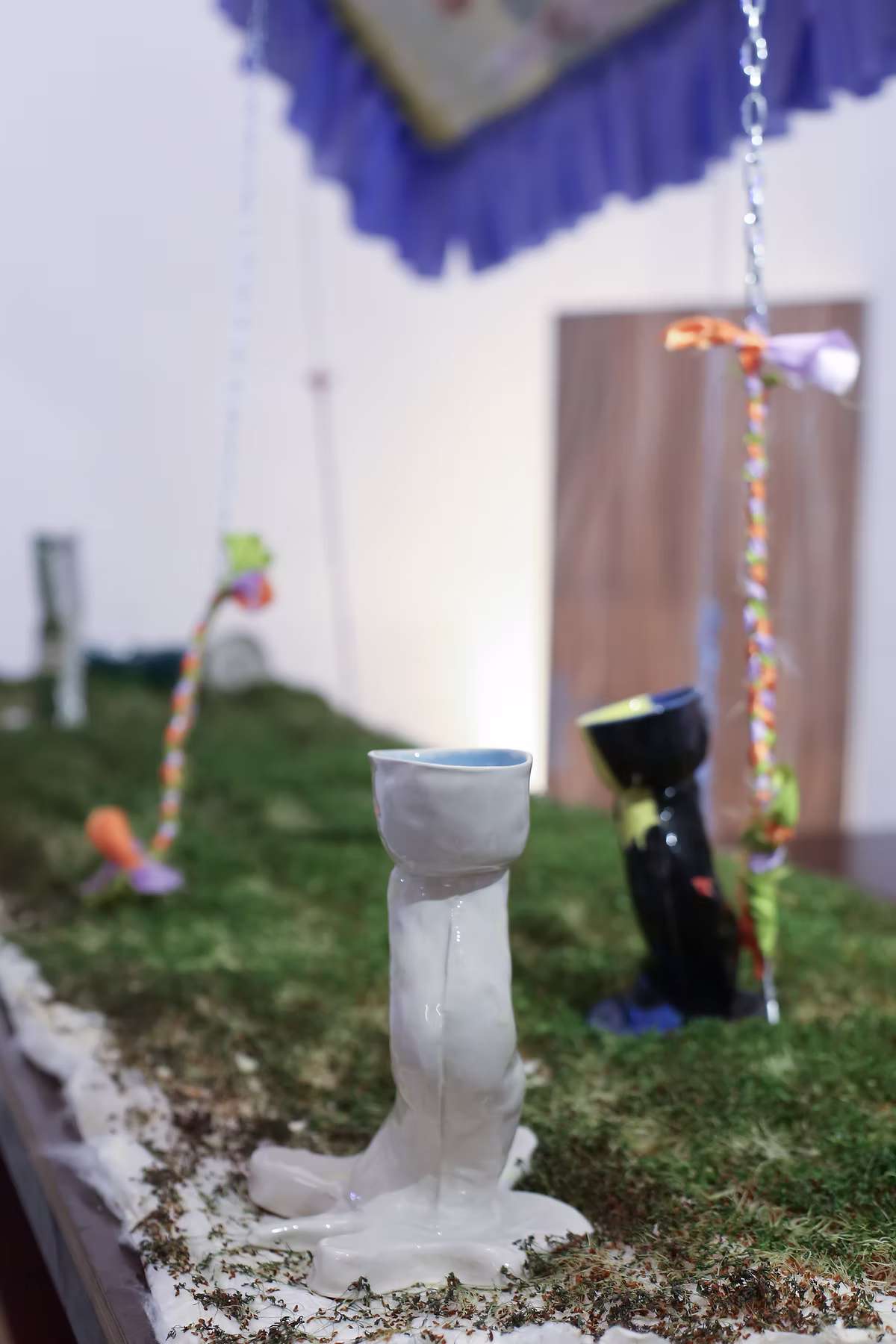A white ceramic vessel on grass with a black vessel and hanging ropes behind it.