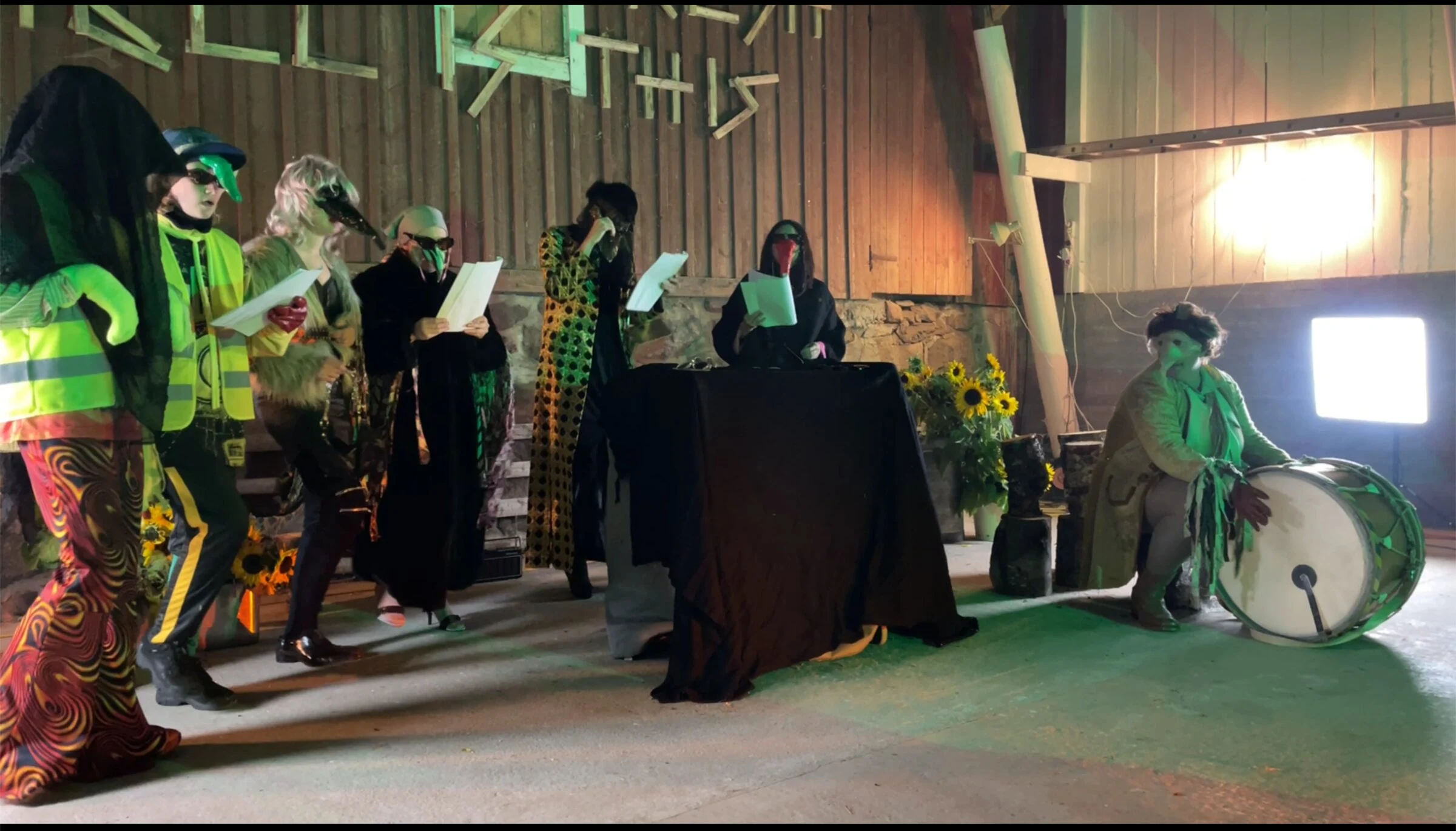 The masked cast reading scripts beside a drum under green stage lighting.