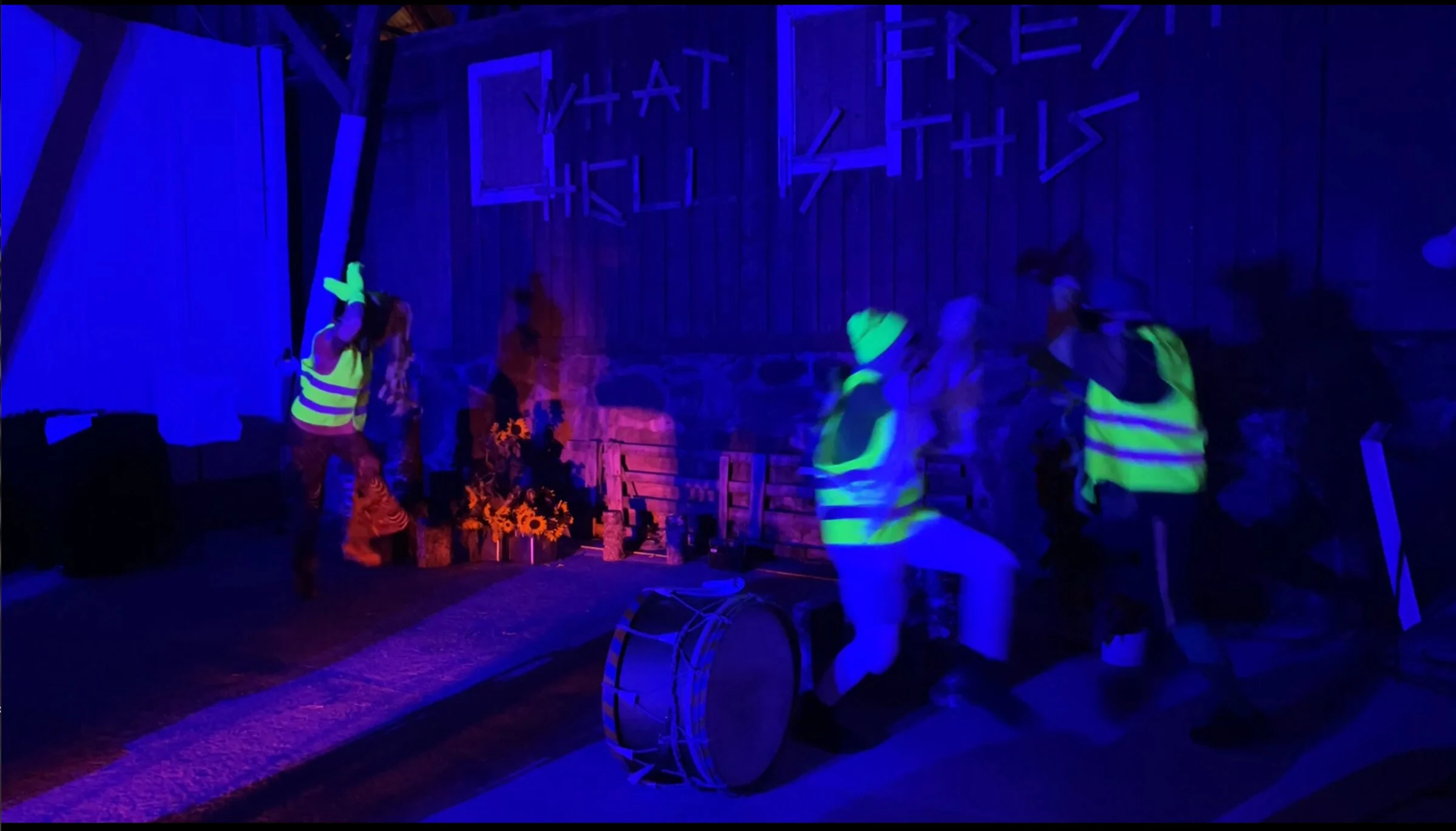 Performers in fluorescent vests dancing beside a drum under blue and purple lighting.