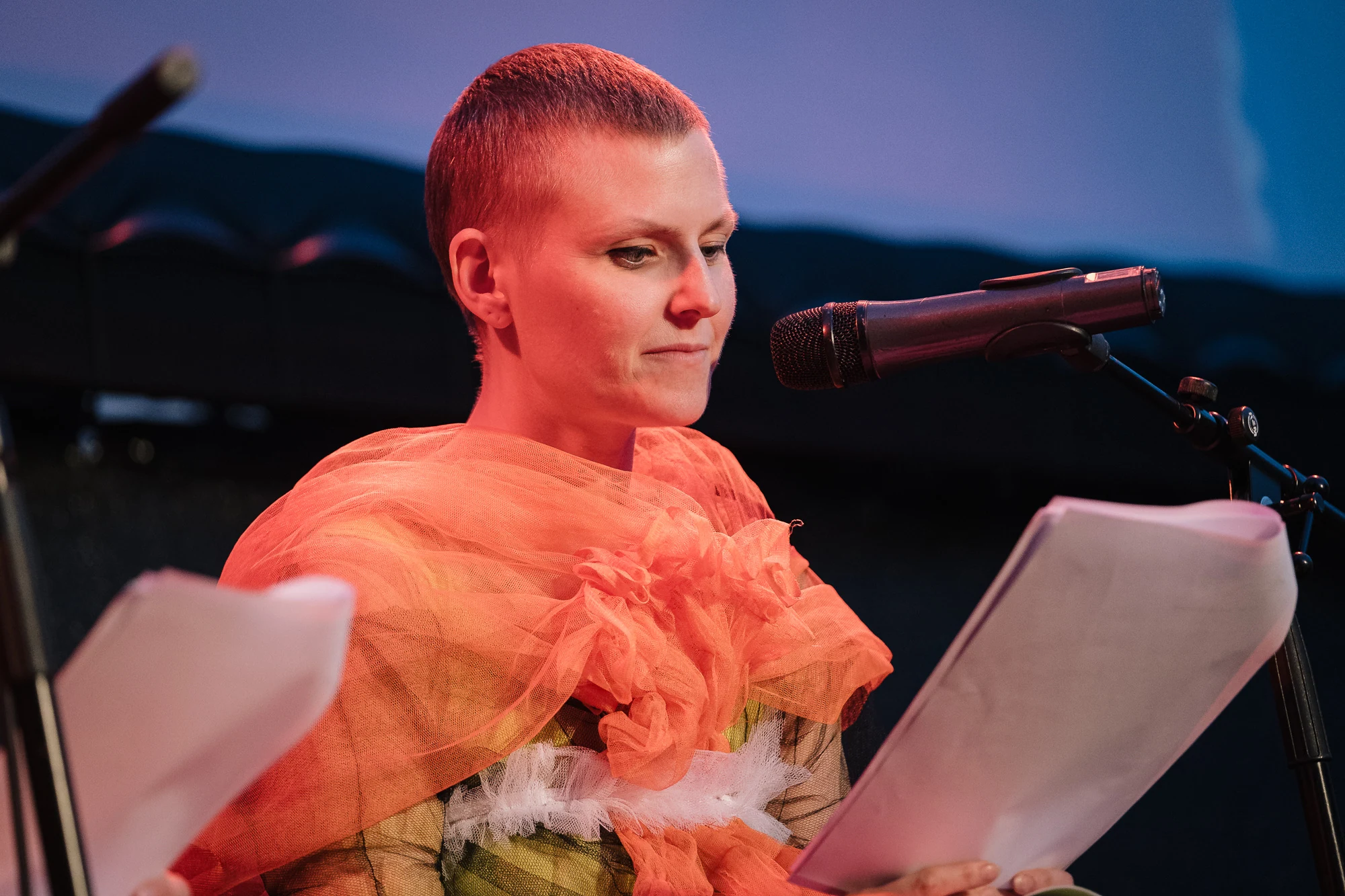 A performer in an orange tulle collar reading a script into a microphone.