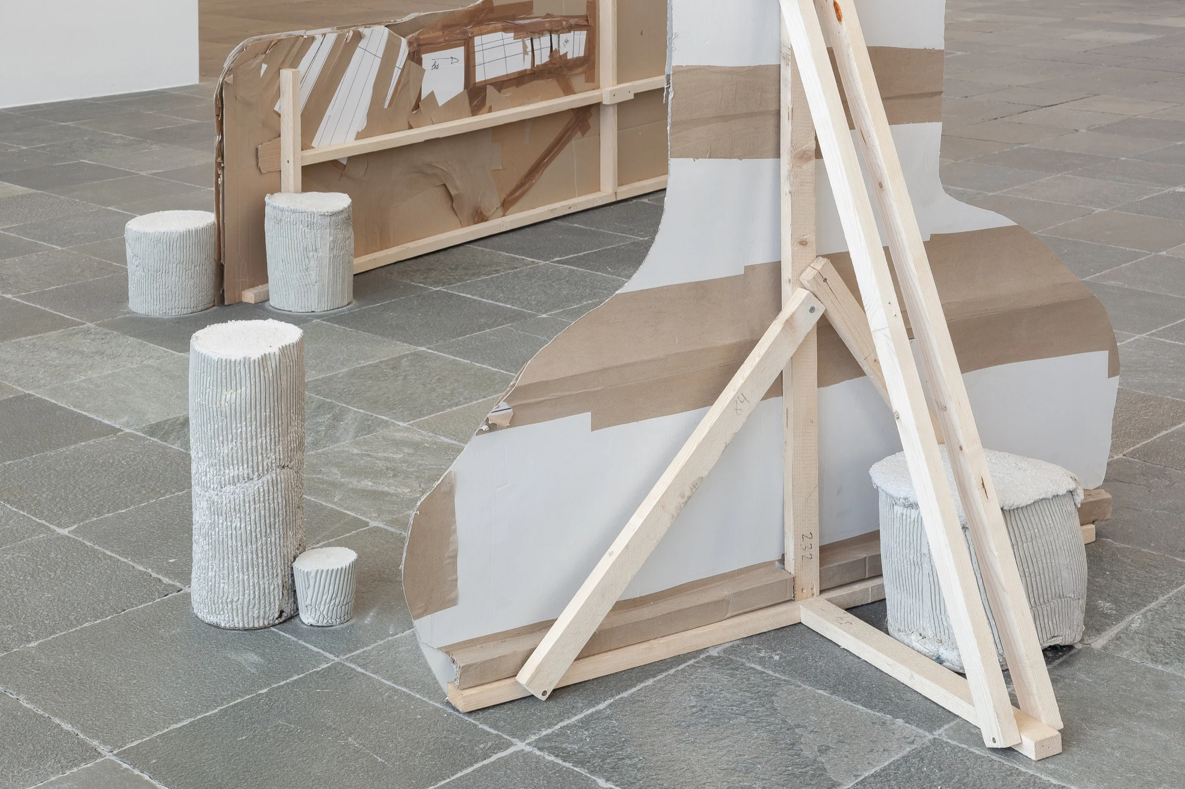 Wooden braces behind the cardboard sculpture, surrounded by white cylindrical objects.