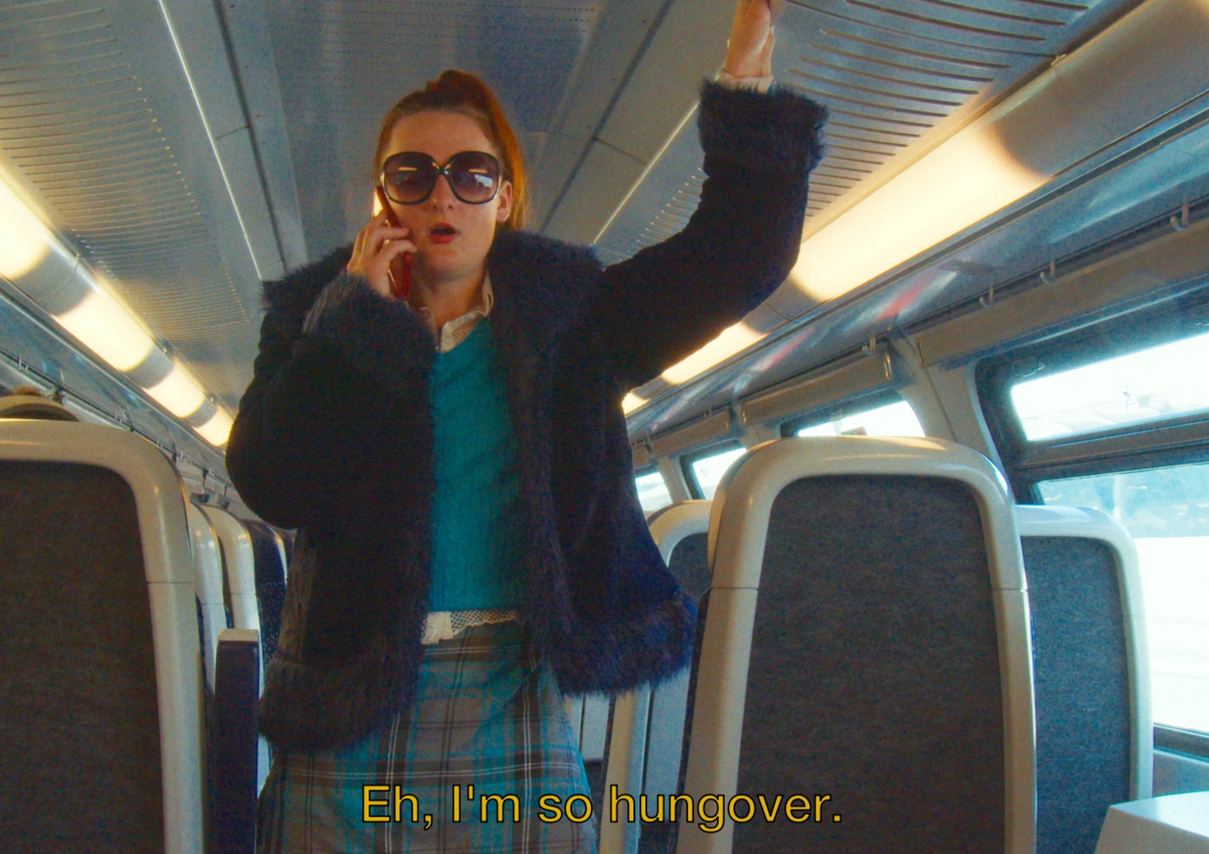 Charlotte wearing sunglasses and talking on the phone on a train, subtitled Eh, I'm so hungover.