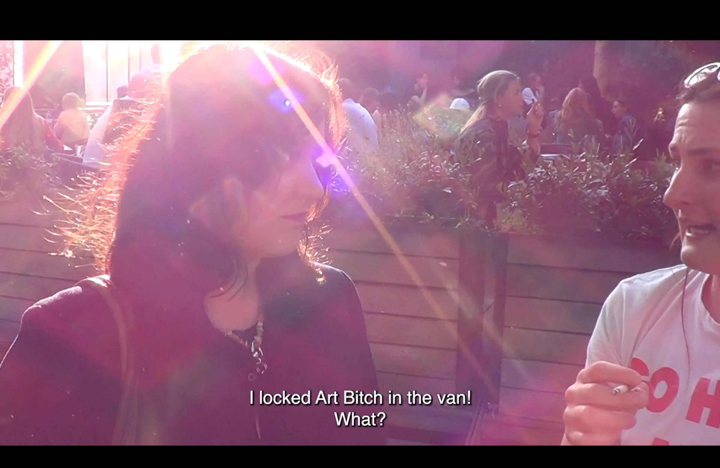 Two characters talking at an outdoor café in bright sunlight, with a subtitle about locking Art Bitch in the van.