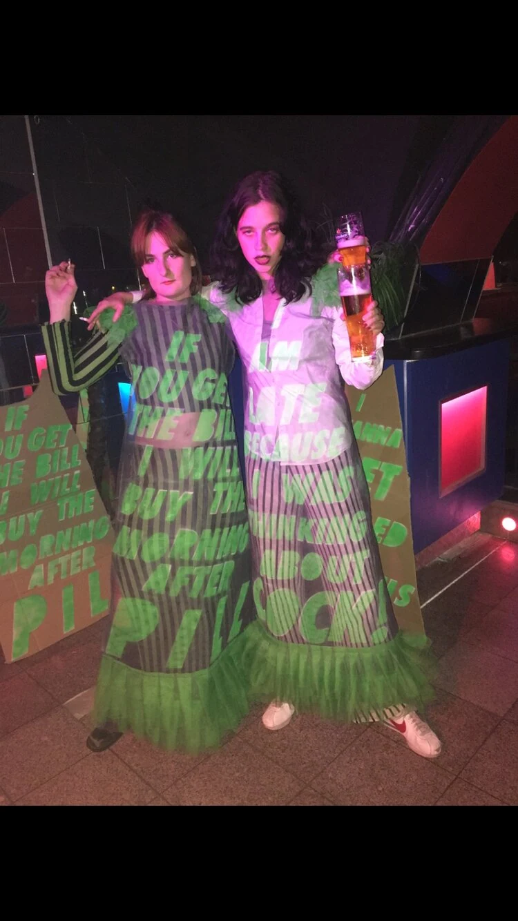 Two performers in dresses with projected green lettering holding beer glasses under pink light.