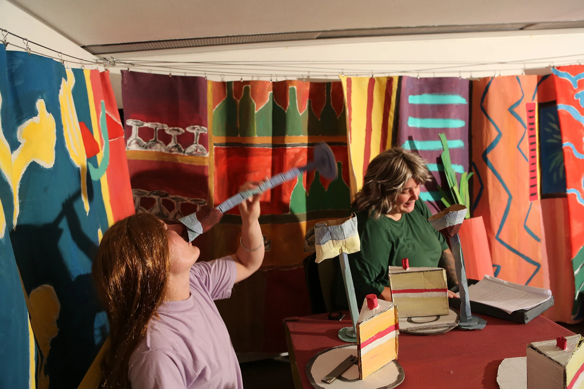 Two performers in wigs seated at a red cardboard table with cake slices and oversized wine glasses.