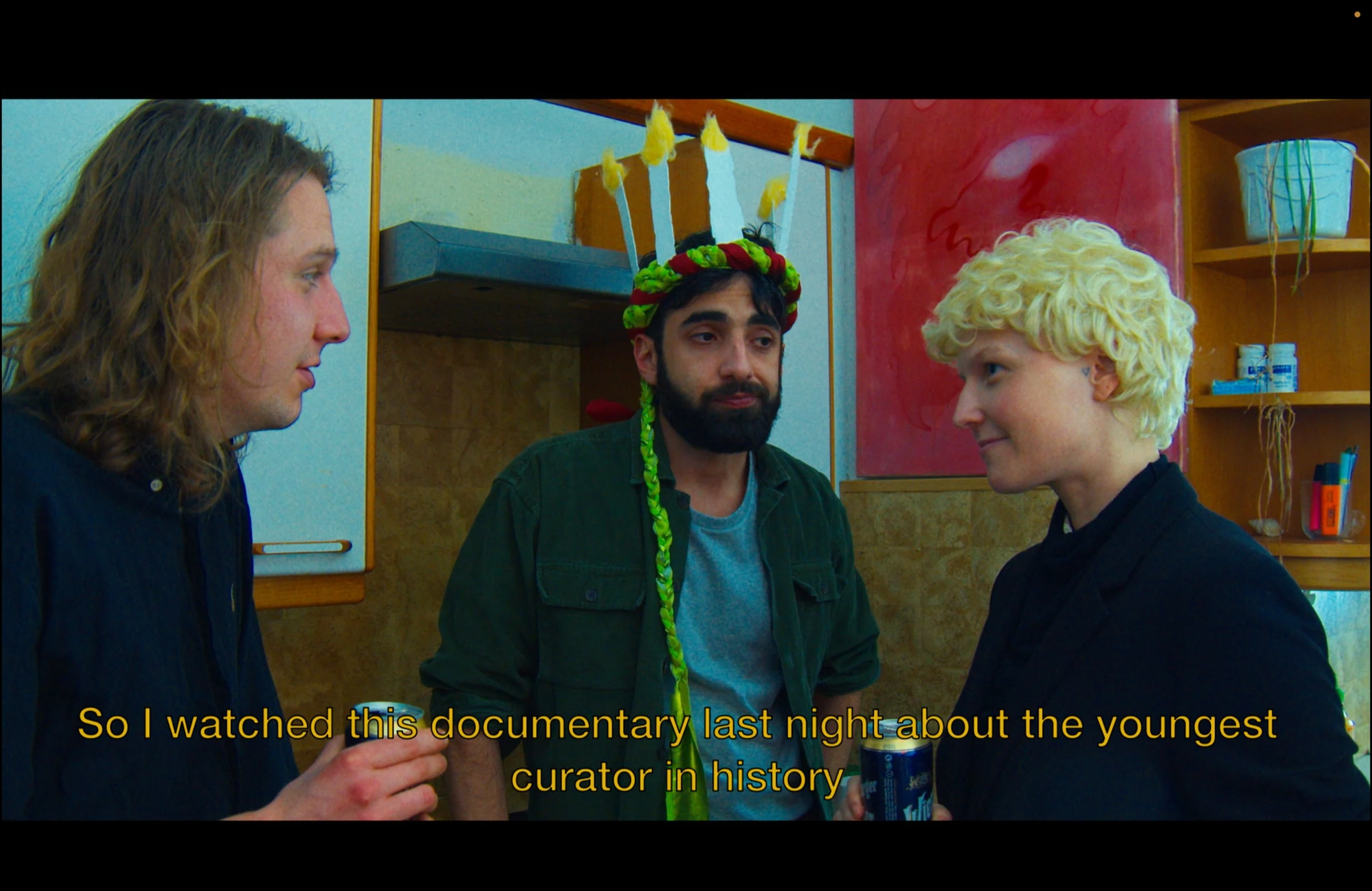 Three friends talking in a kitchen, one wearing a crown of handmade candles, with a subtitle about the youngest curator in history.